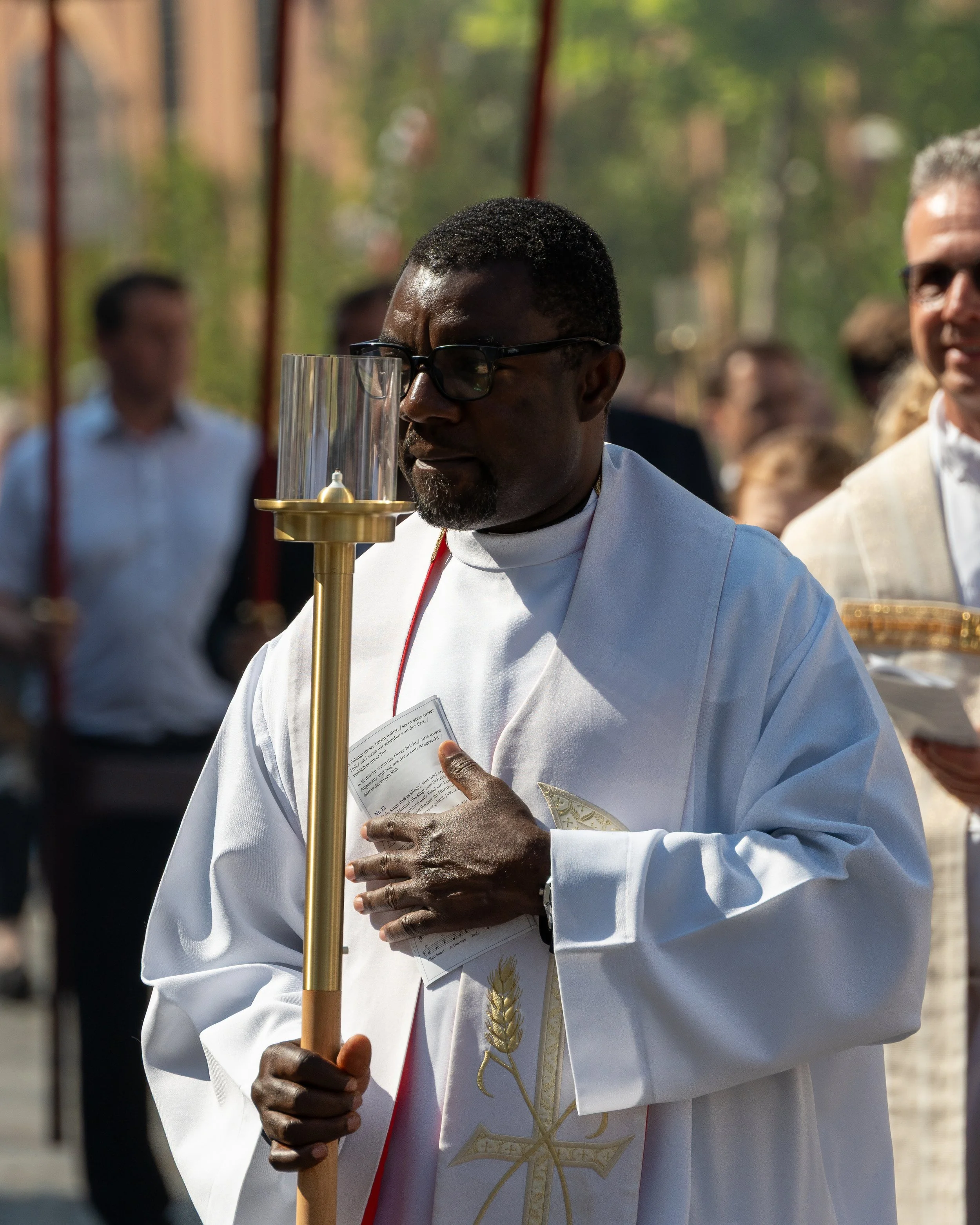 A priest holding a large candle during a religious ceremony outdoors, surrounded by other attendees.