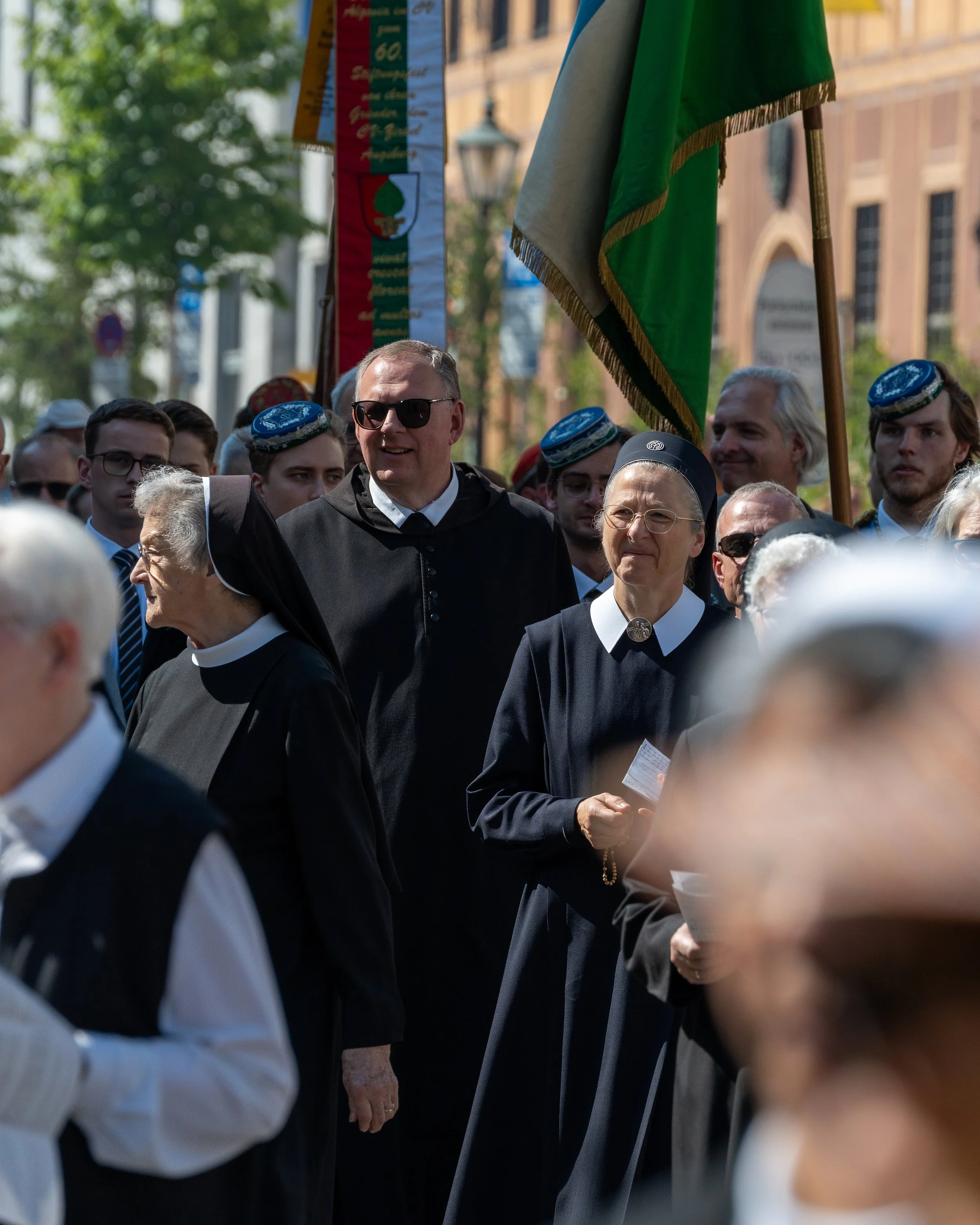 A crowd participating in a parade or march, including nuns, a priest, and people holding flags, with trees and building background.