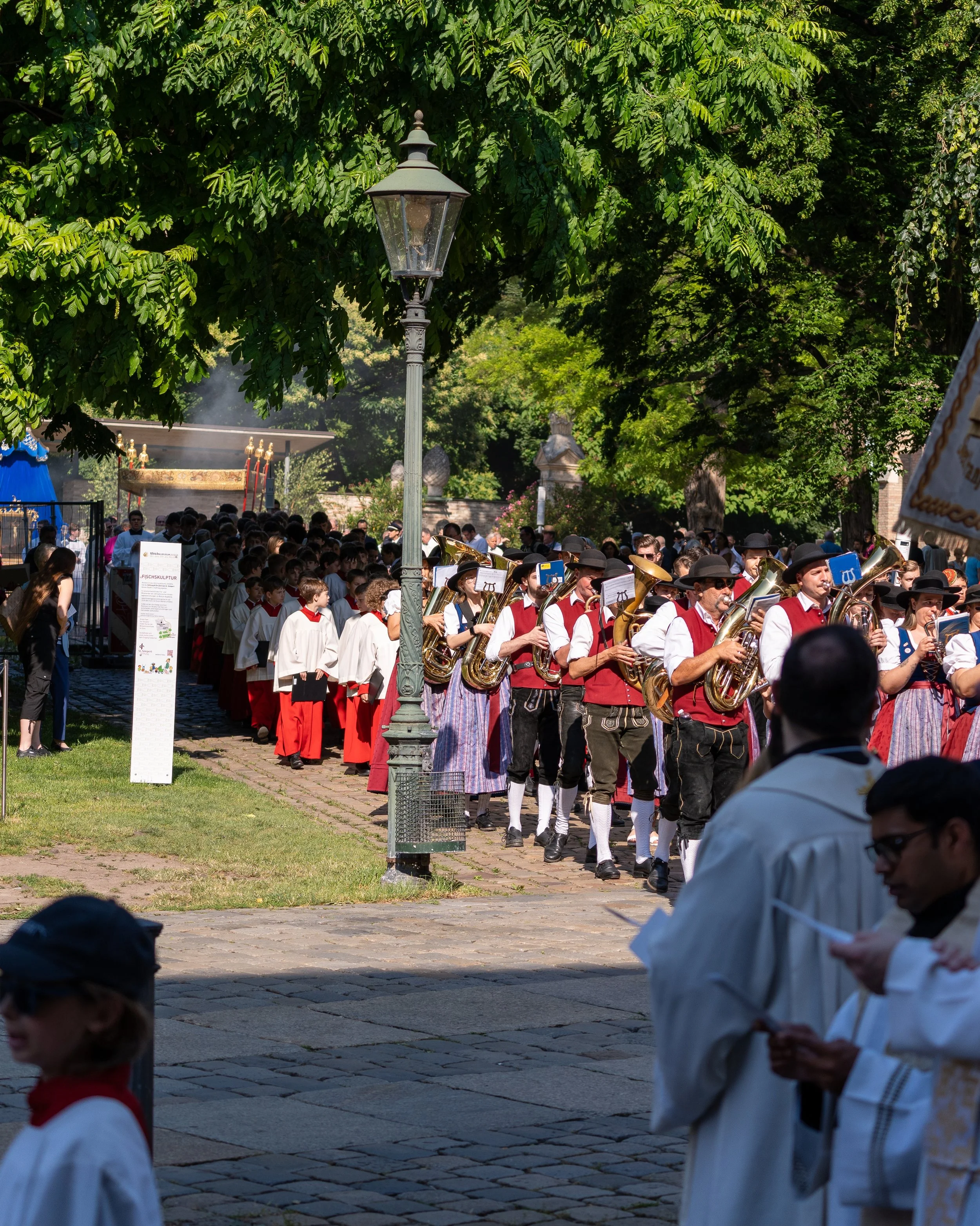 A parade with a band of musicians in traditional Bavarian attire walking on a cobblestone path, surrounded by a crowd and lush green trees on a sunny day.