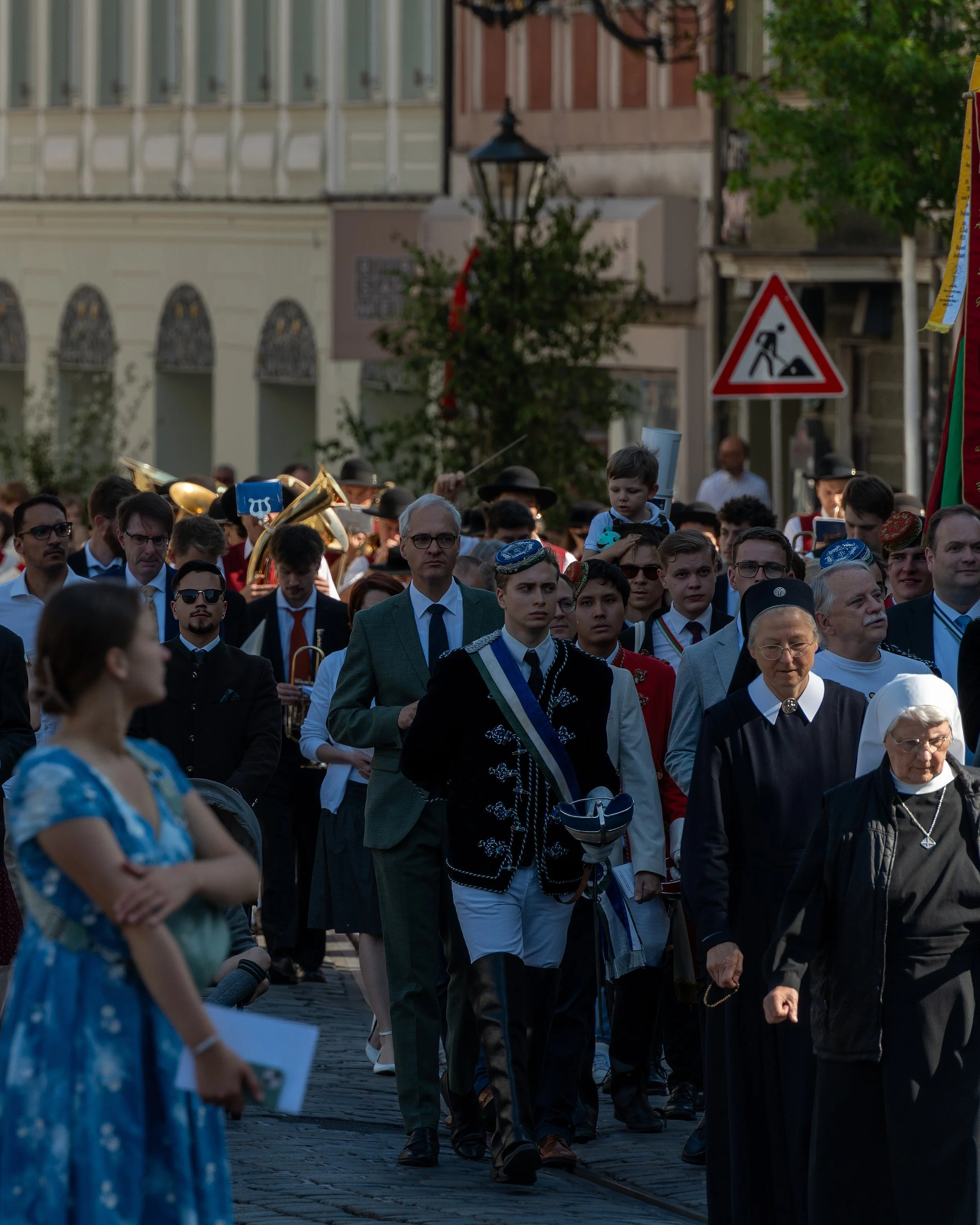 People marching in a street parade, including children and adults dressed in various costumes and uniforms, with musicians playing brass instruments in the background, amidst city buildings and greenery.
