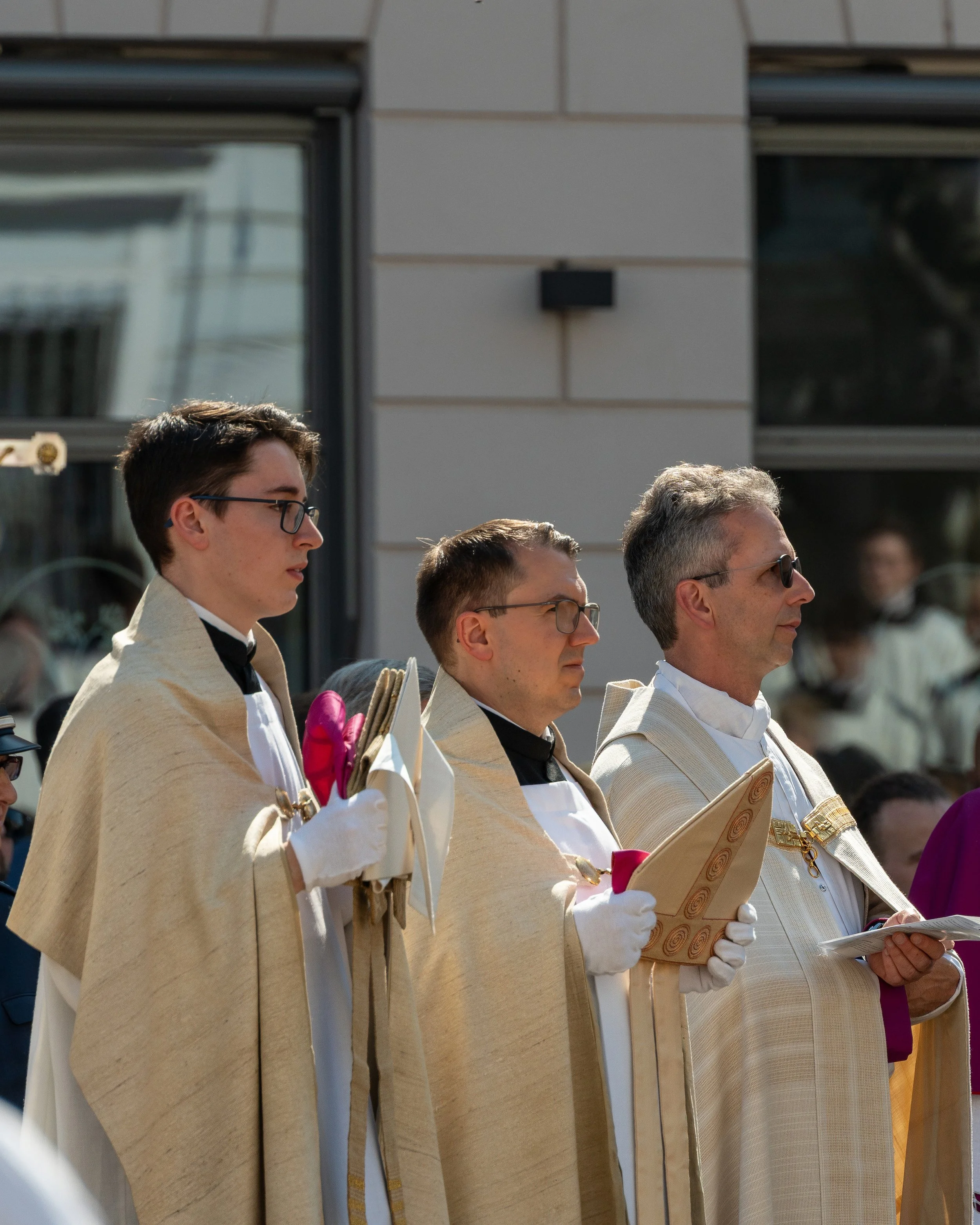 Three priests dressed in light-colored robes participating in a religious procession, holding religious items, outdoors during daytime.
