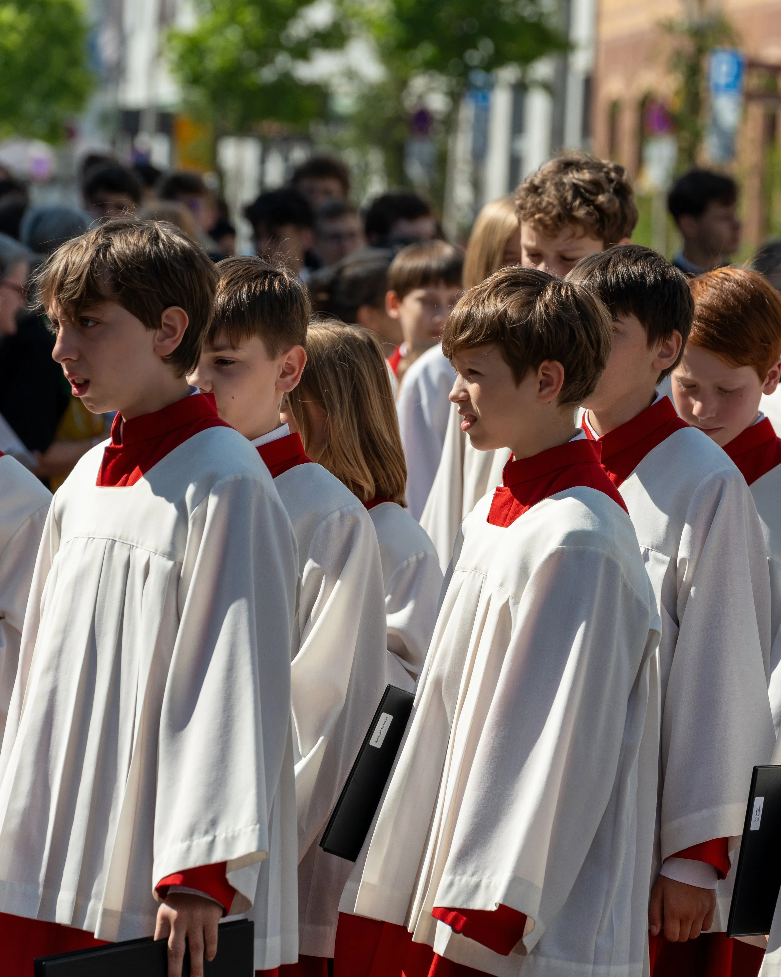 Group of children dressed in choir robes standing outdoors during daytime, holding black folders, with trees and buildings in the background.