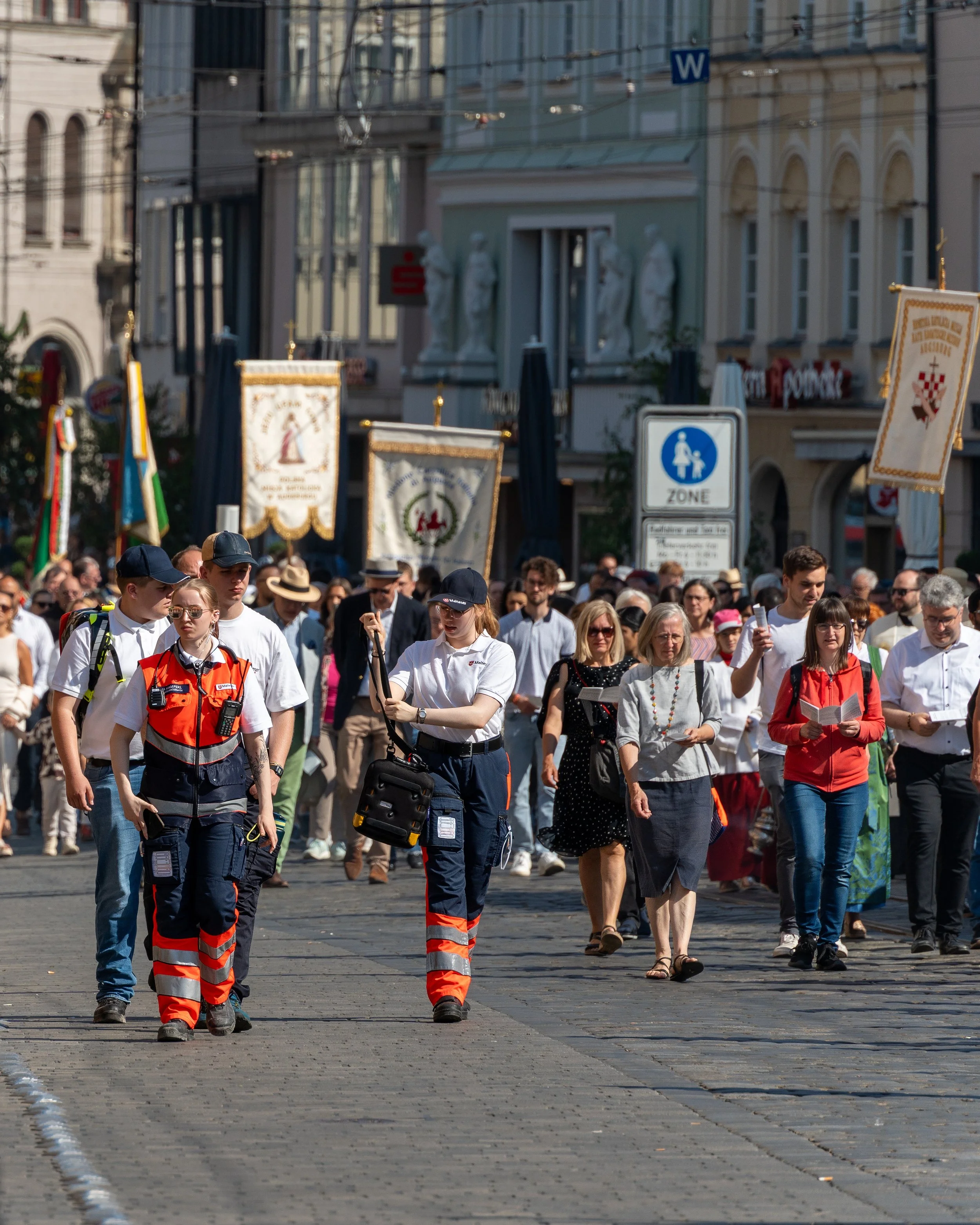 A large crowd of people walking on a city street, with some holding banners, and two emergency medical workers in orange and navy uniforms leading the way.