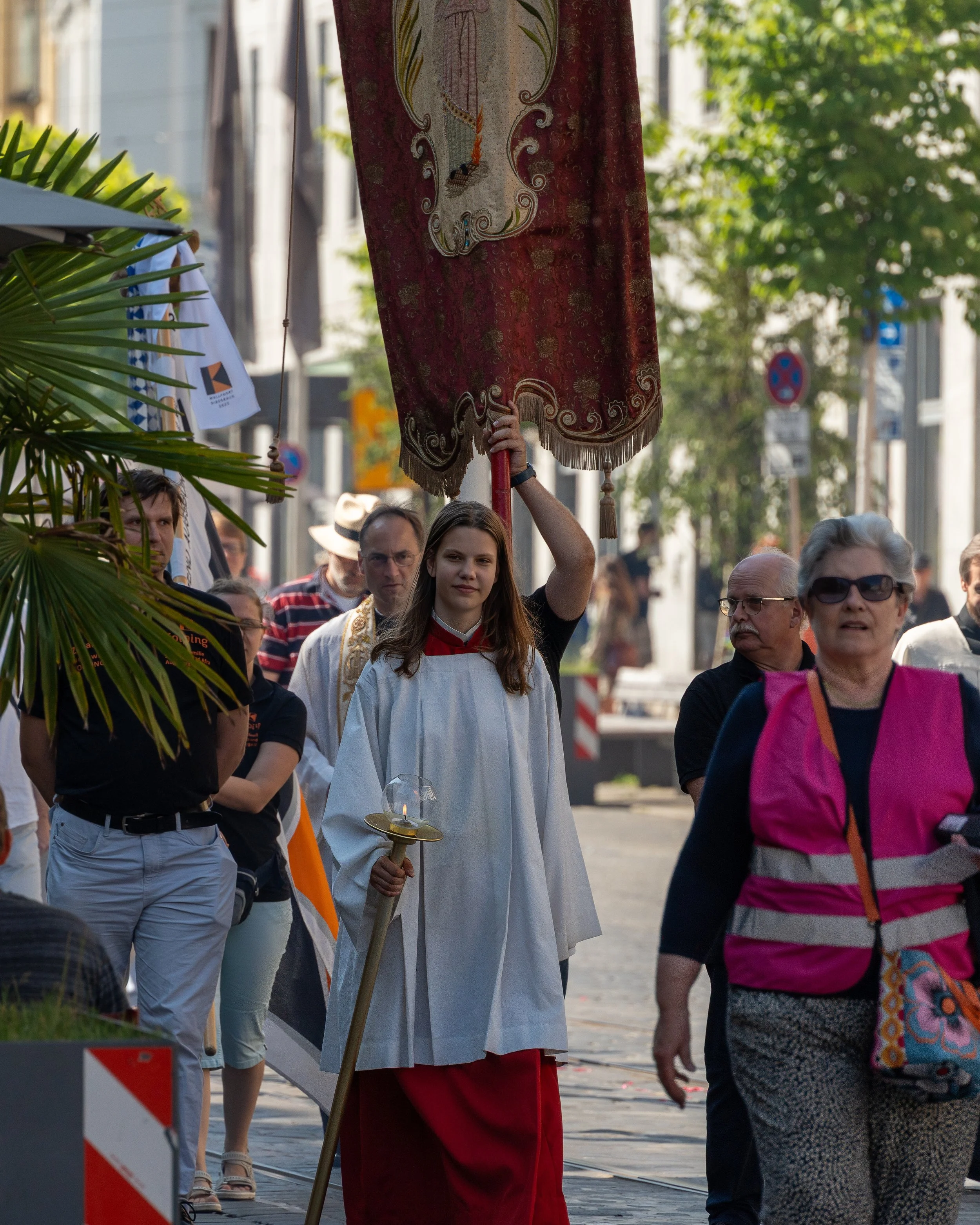 A young woman in a white altar server robe carrying a liturgical thurible and a large red ceremonial banner leading a religious procession through a city street, with onlookers and trees in the background.