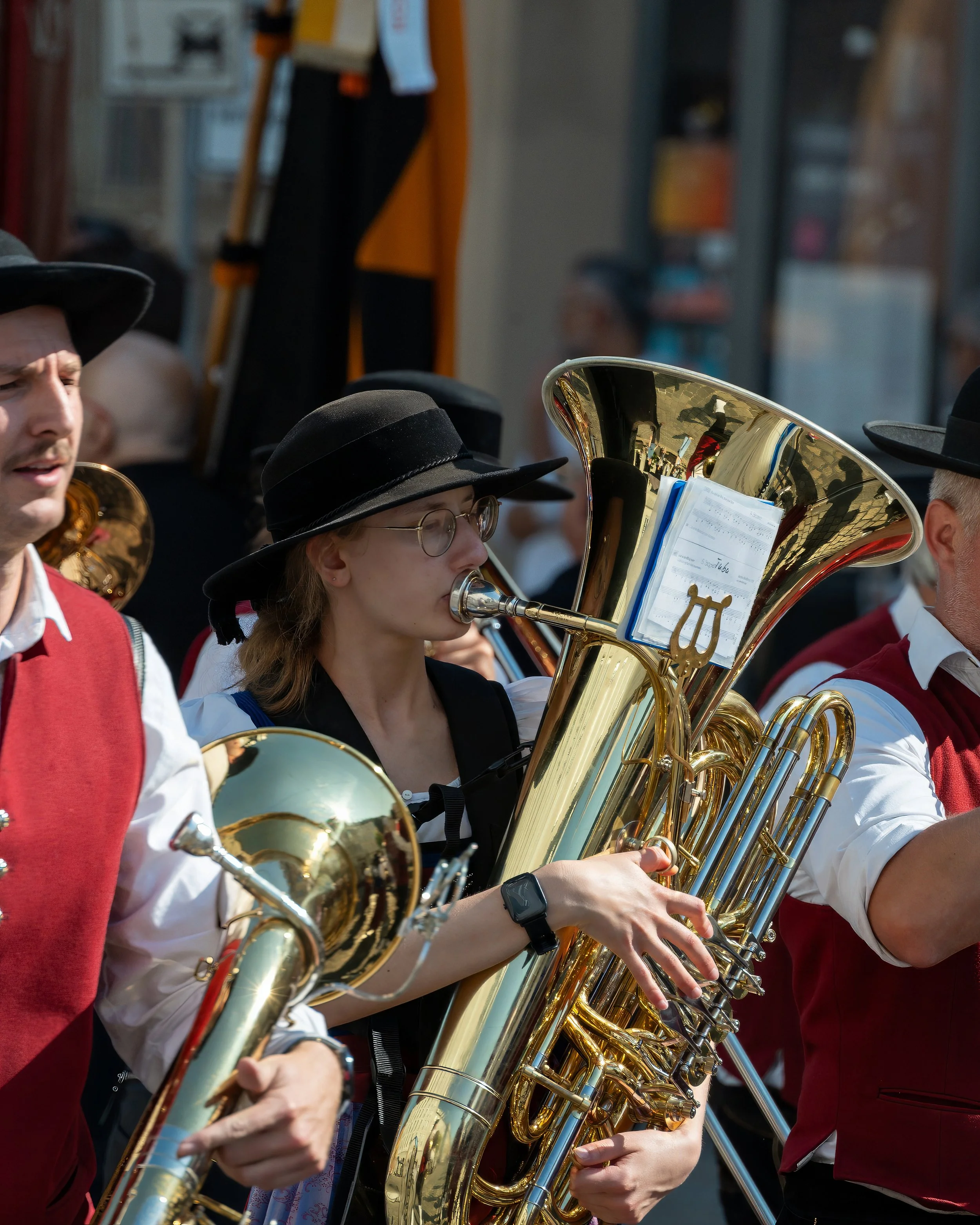 A woman playing a brass sousaphone in a marching band, wearing glasses, a black hat, and traditional attire, surrounded by other band members also in traditional uniforms.
