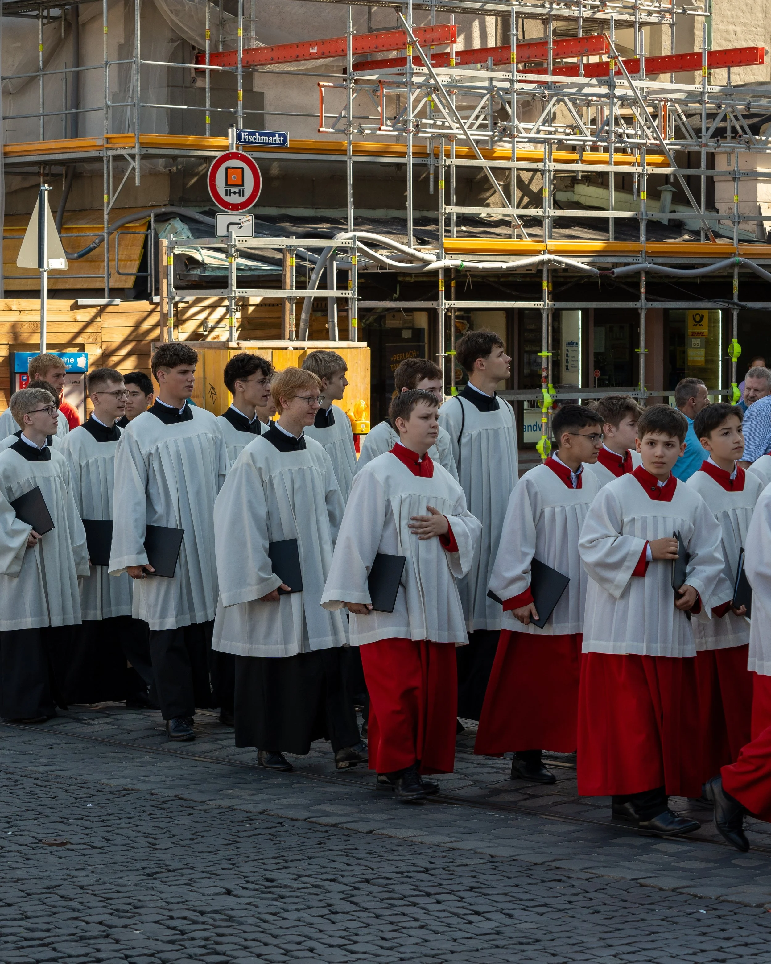 Group of people dressed in choir robes, participating in a procession on a cobblestone street with construction scaffolding in the background.