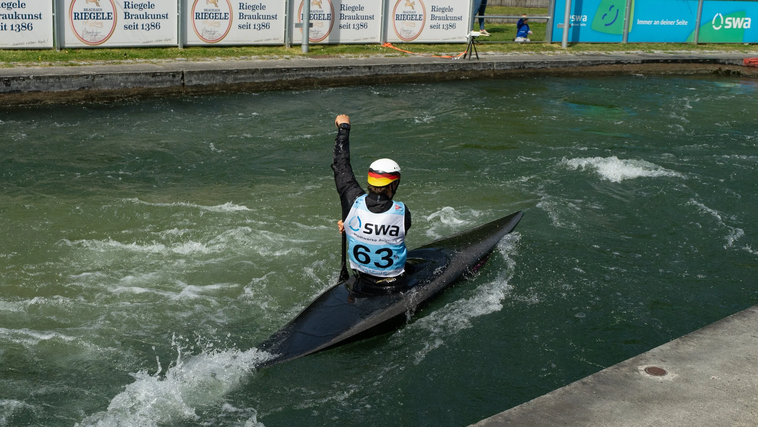 A kayaker wearing a white helmet with a German flag design, a black jacket, and a vest with the Swa logo and the number 63, raising their right fist while sitting in a black kayak in a water channel during a race.