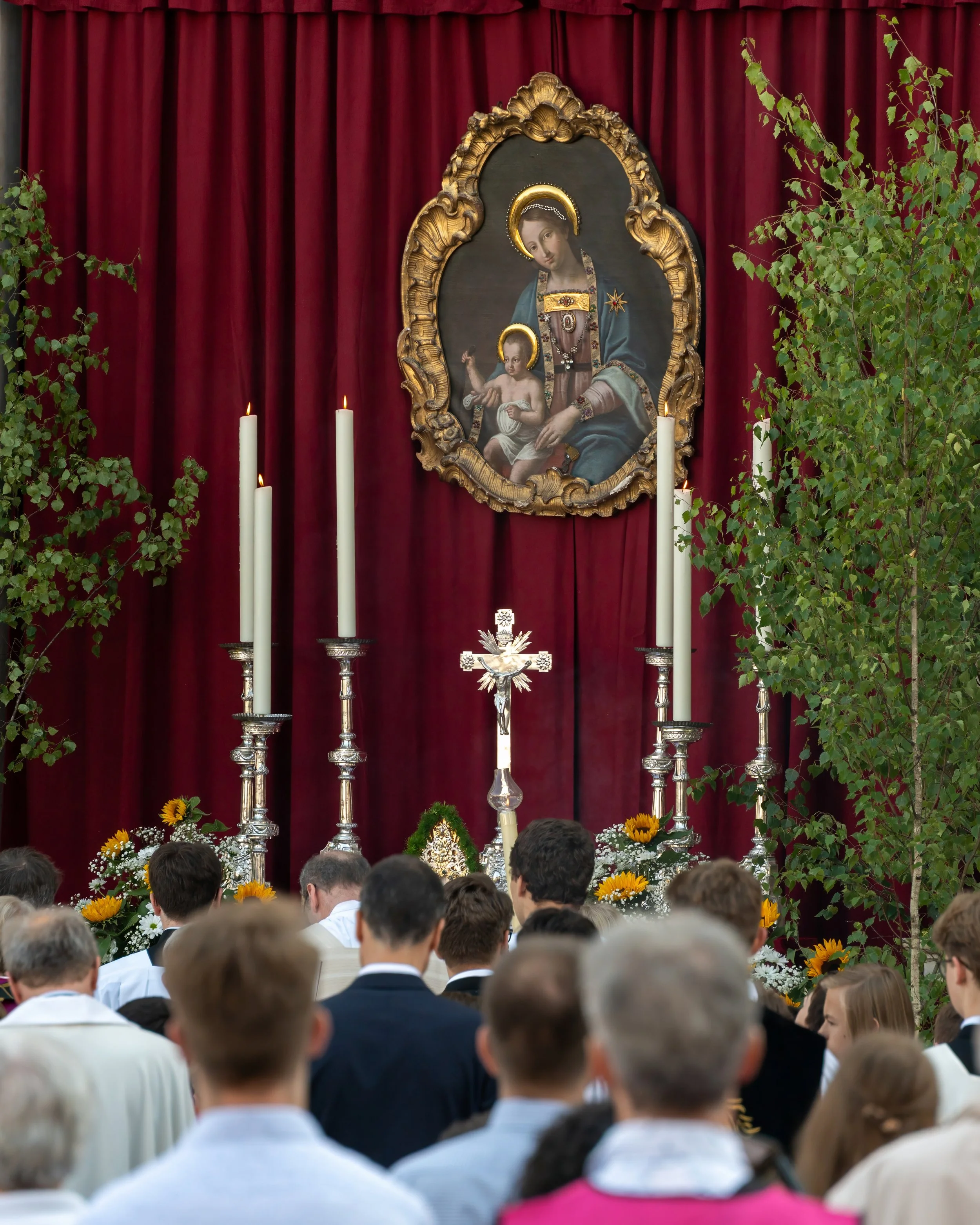 People gathered in front of a religious altar with candles, flowers, and a crucifix, featuring a large framed painting of the Virgin Mary holding baby Jesus, with red curtains and green trees on either side.