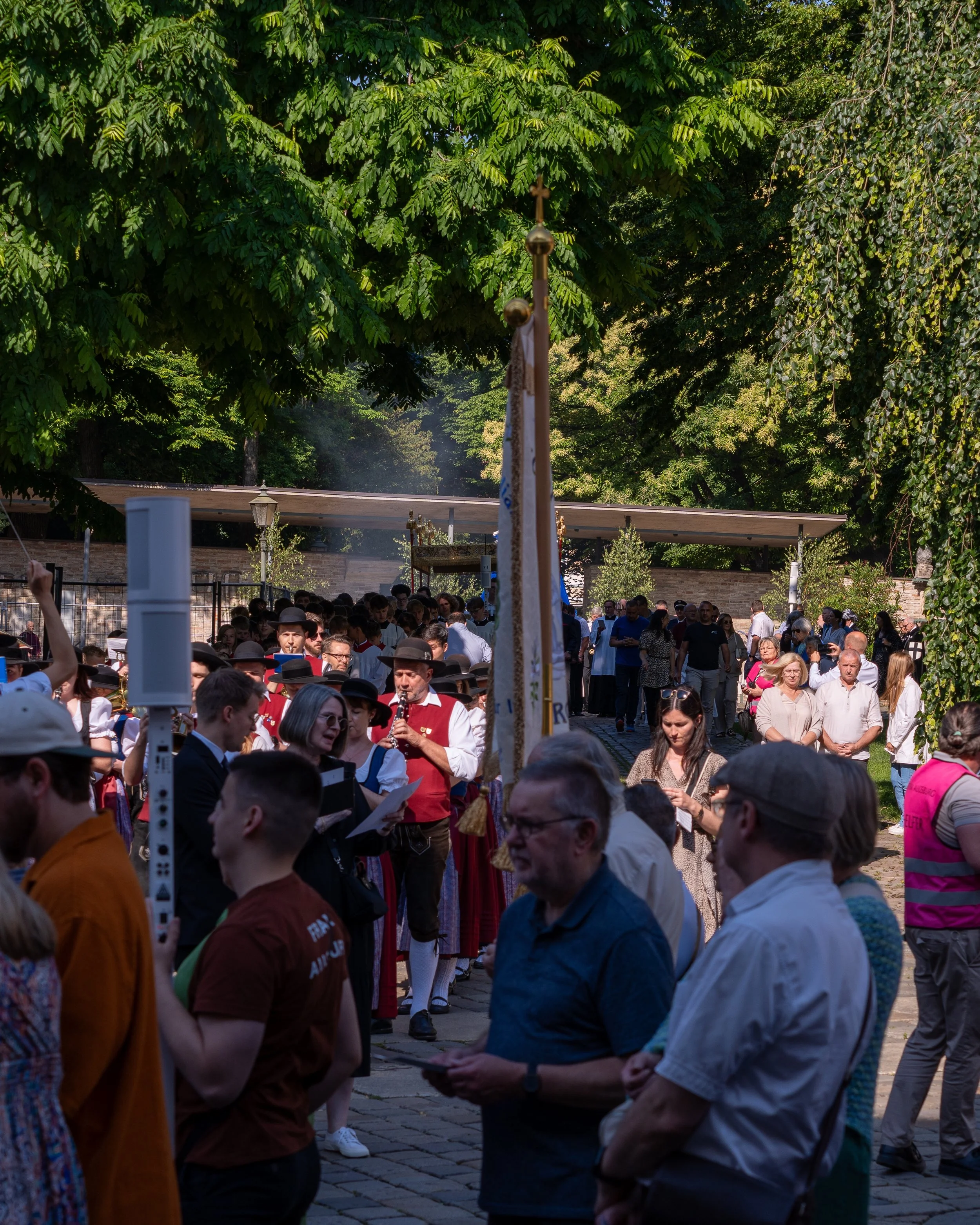 A crowd of people participating in an outdoor cultural or religious event, with some dressed in traditional attire, under trees on a sunny day.