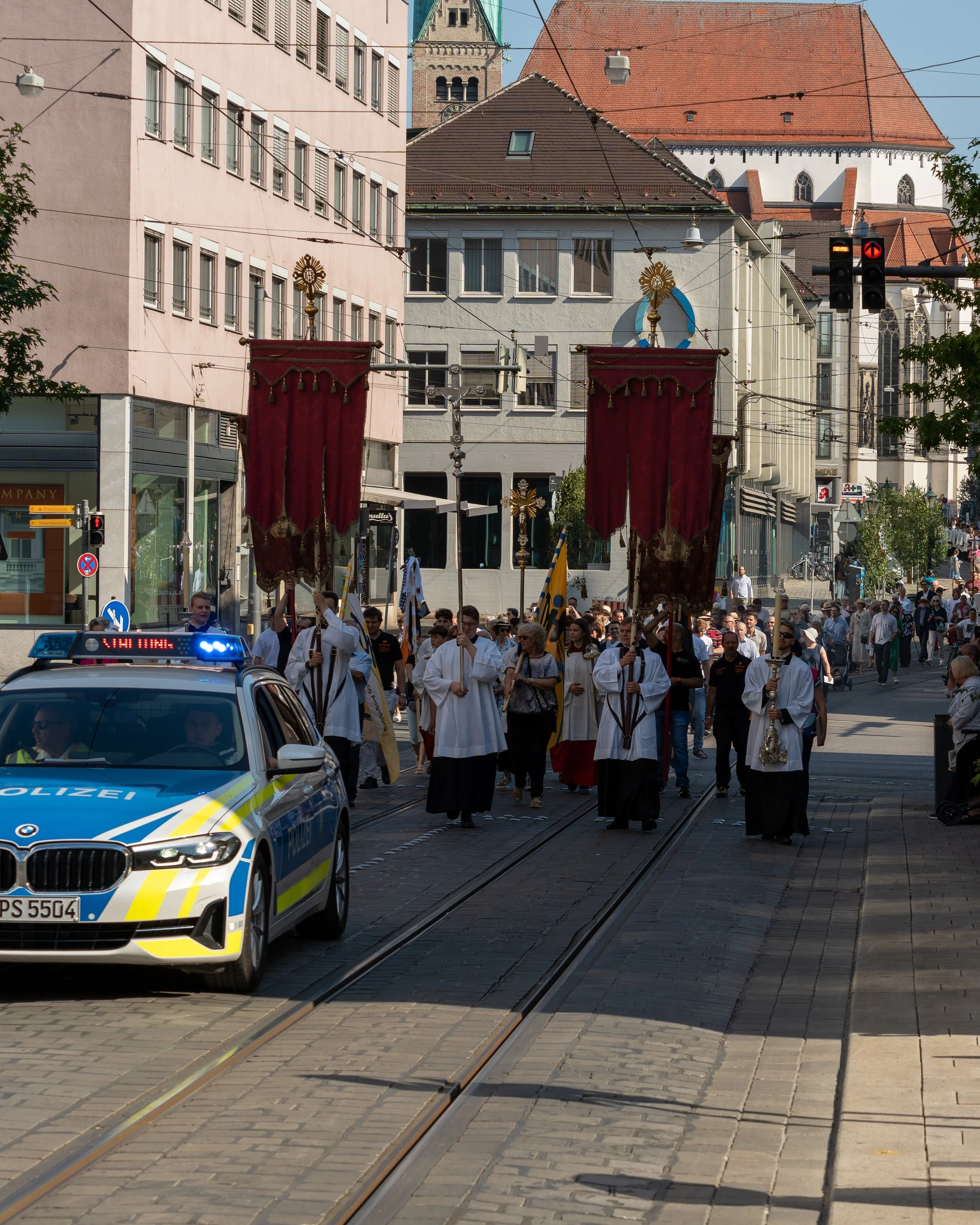 A religious procession with people in white robes carrying banners and crosses, walking along tram tracks in an urban area with buildings, a church, and a police car nearby.