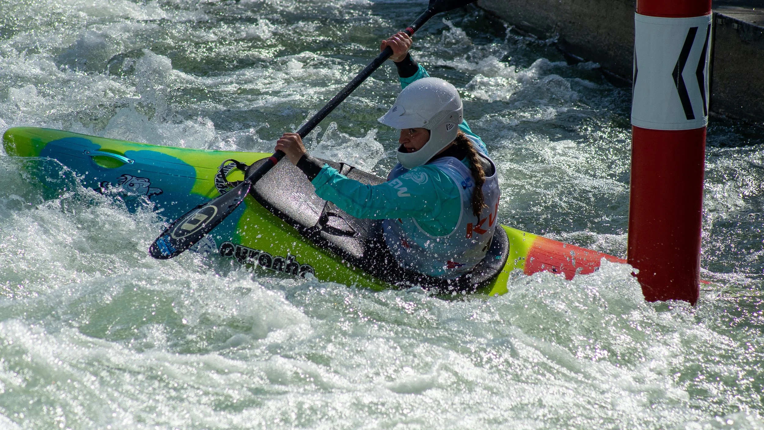 A female kayaker wearing a helmet and a life jacket navigates through white water rapids, paddling past a red and white gate on the river.