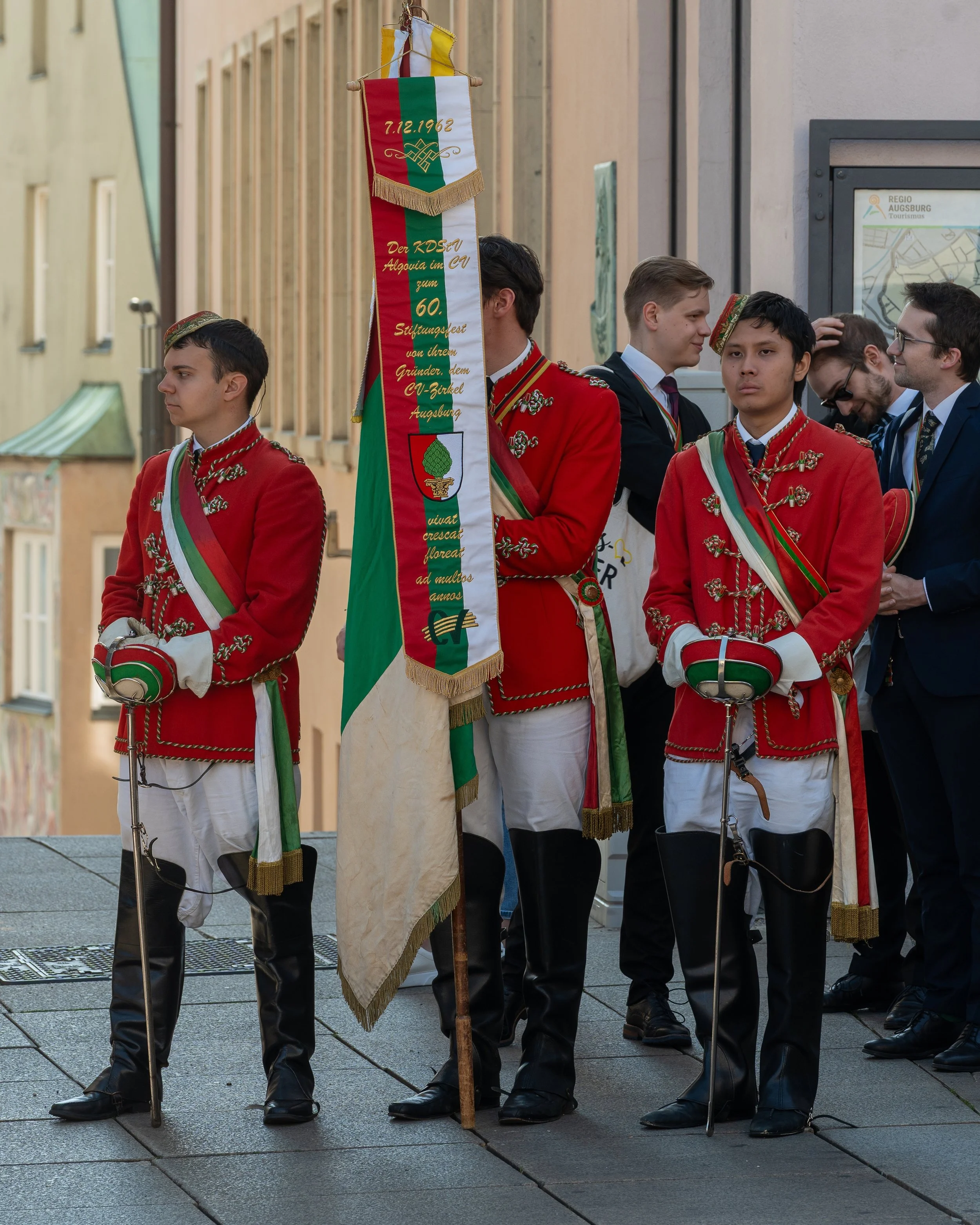 Young men dressed in traditional red and white uniforms, holding a flag with green, white, and red stripes, standing in a line during a parade or ceremony.