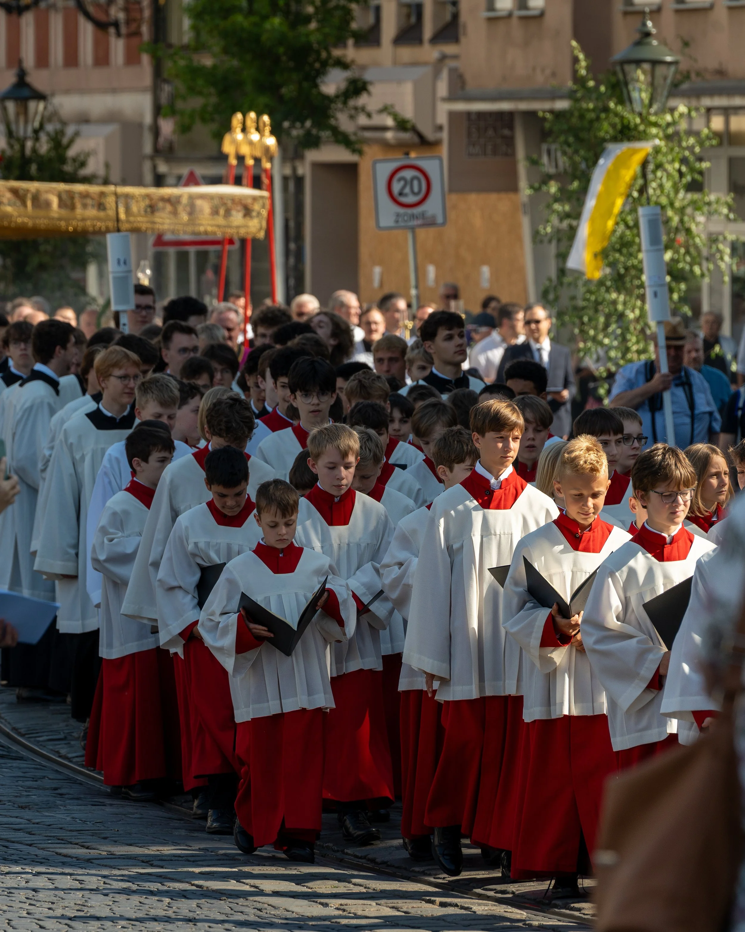 A group of children dressed in choir robes, walking in a procession. They are holding black hymnals, and the procession is taking place on a cobblestone street with onlookers and decorations in the background.