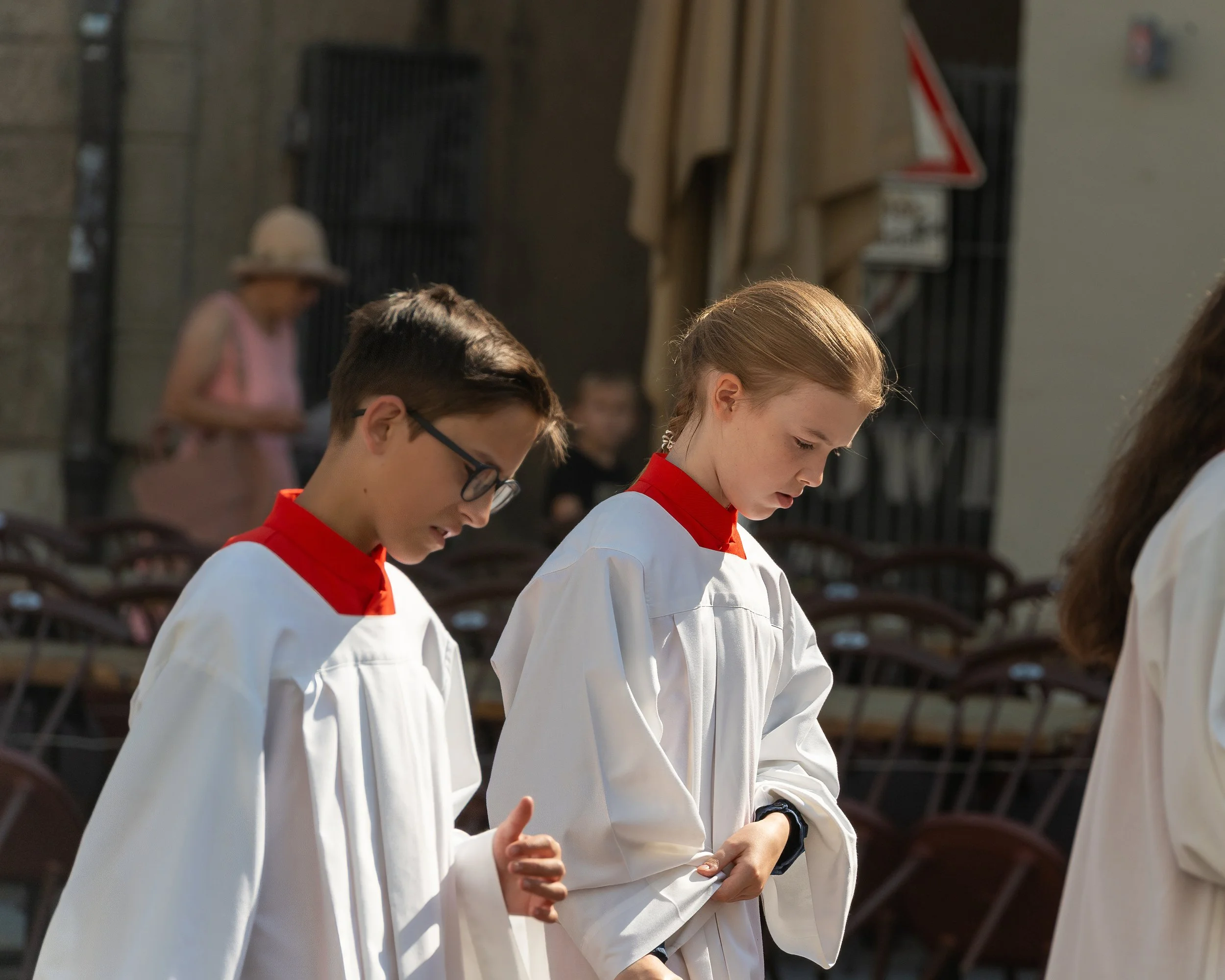 Two children dressed in white robes with red collars praying outdoors.