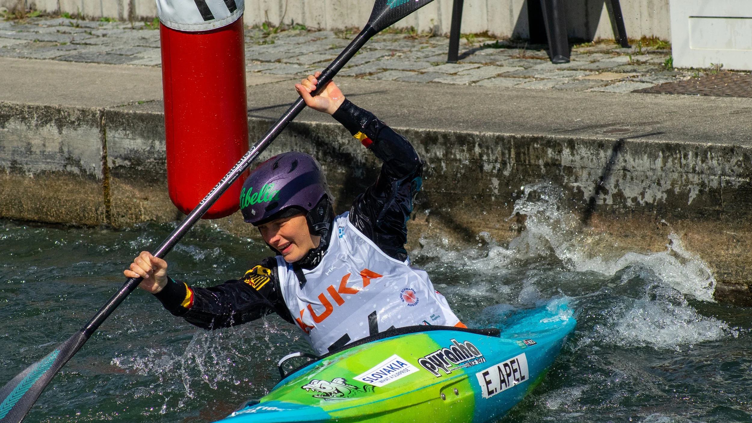 A person wearing a purple helmet and black wetsuit paddles a kayak in a river near a concrete dock, splashing water as they maneuver.