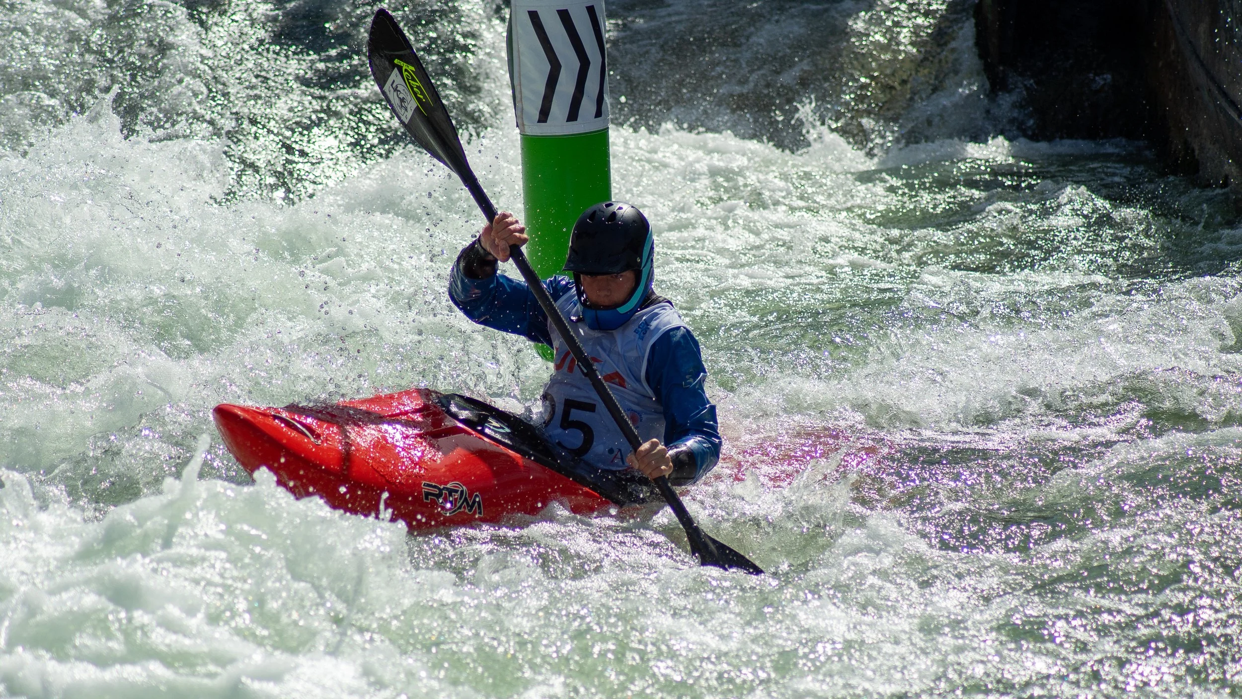 A kayaker wearing a helmet and life jacket navigates white water rapids, holding a paddle above their head, near a green and white marker in turbulent water.