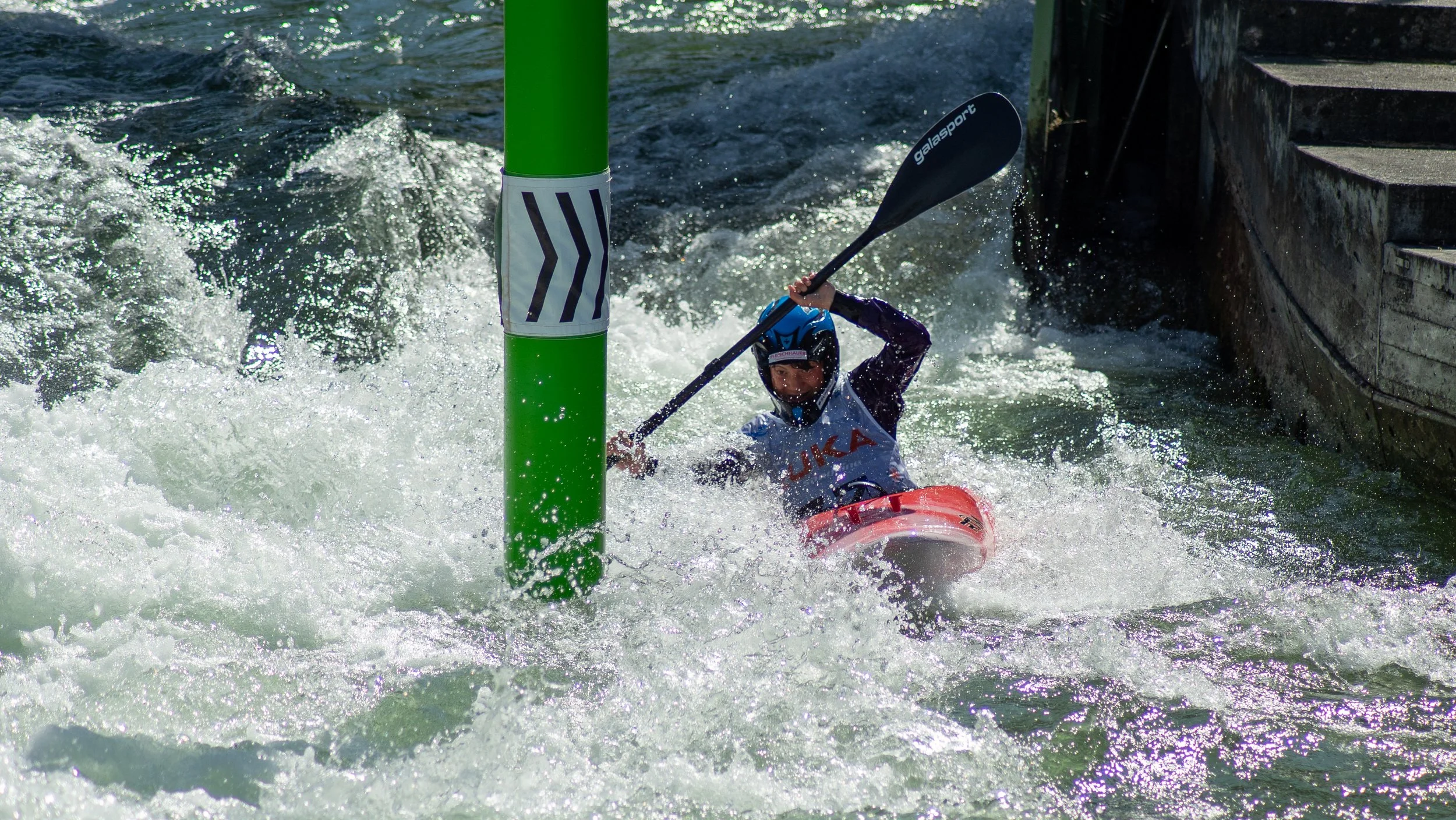 A person kayaking in white water rapids, wearing a helmet and life vest, navigating past a green pole in the water.