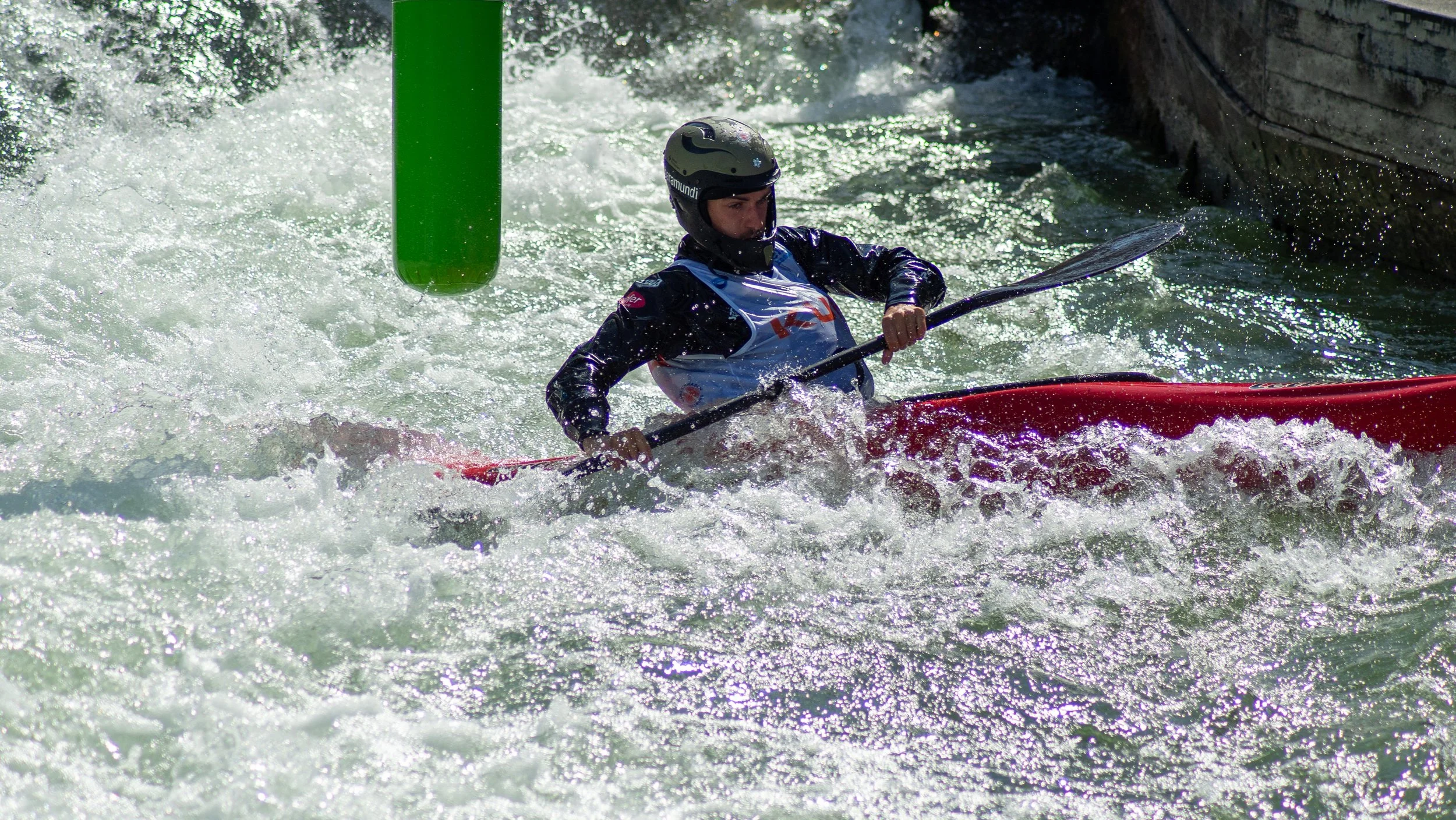 A person wearing a helmet and life jacket paddling a red kayak through white water rapids in a river, near a green buoy and rocks.