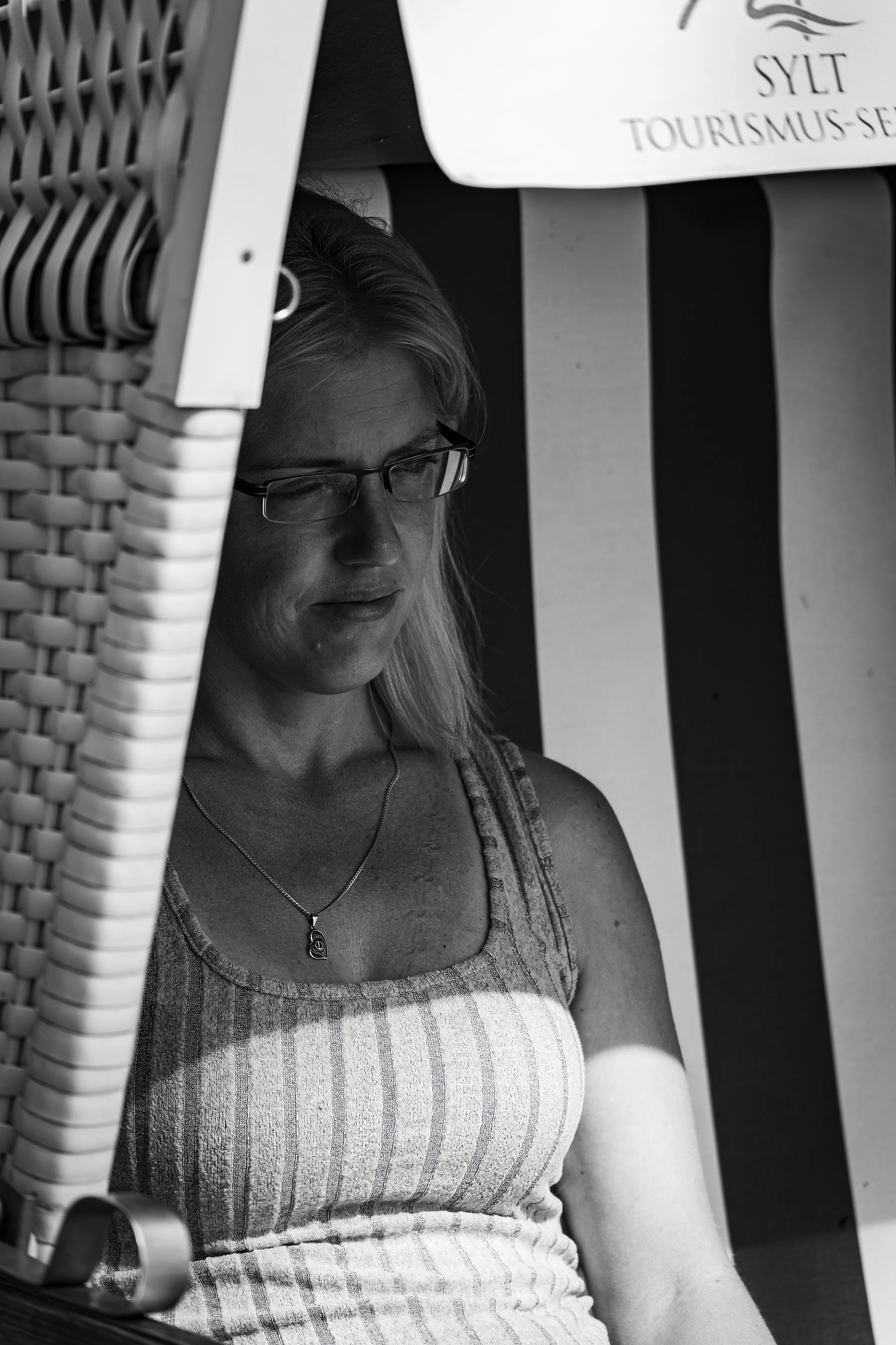 A woman with glasses and a striped tank top sitting outdoors under a striped umbrella, looking down, in black and white.