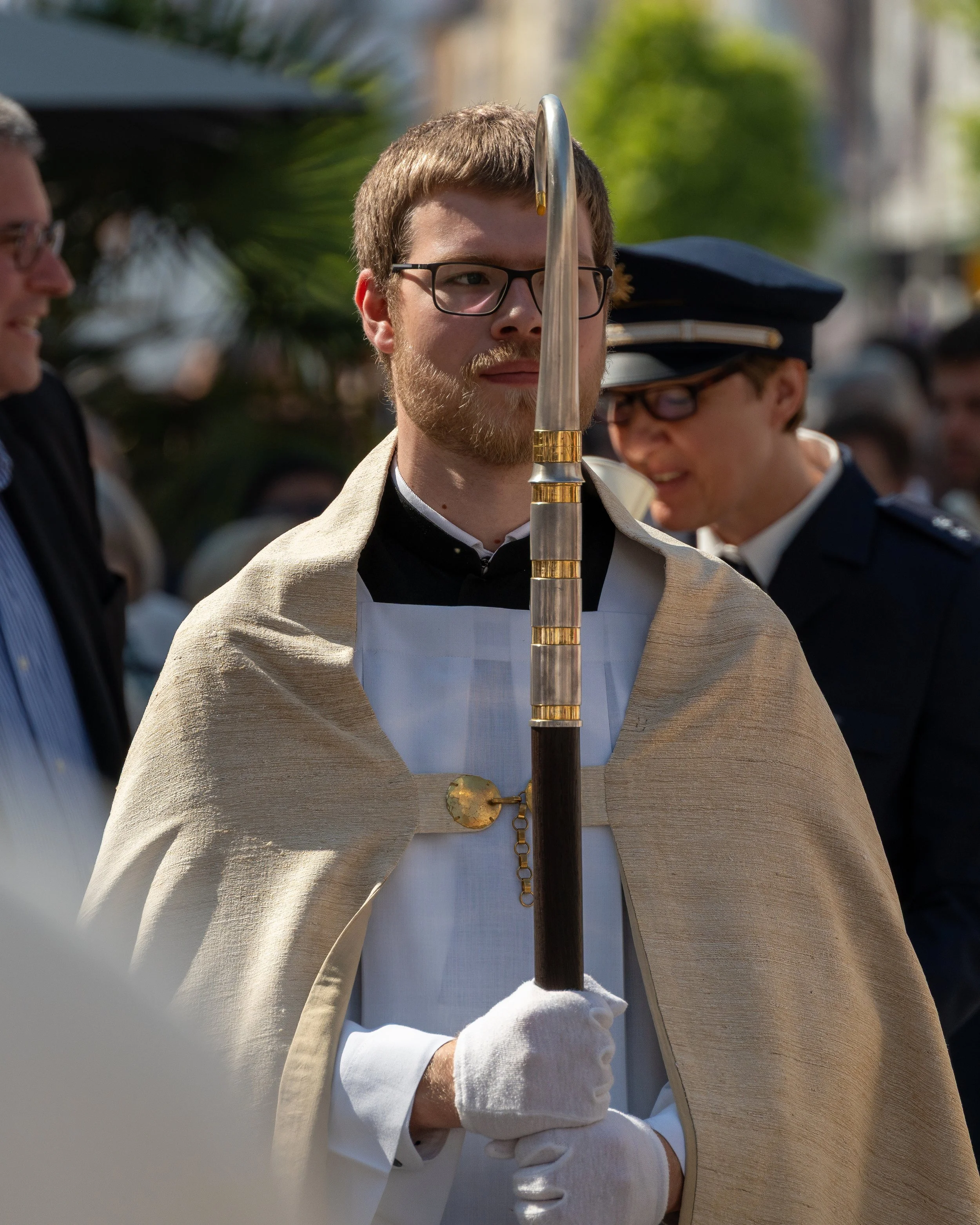 A man dressed in ceremonial robes and gloves holding a pole, participating in a religious procession or event, with several people in the background.