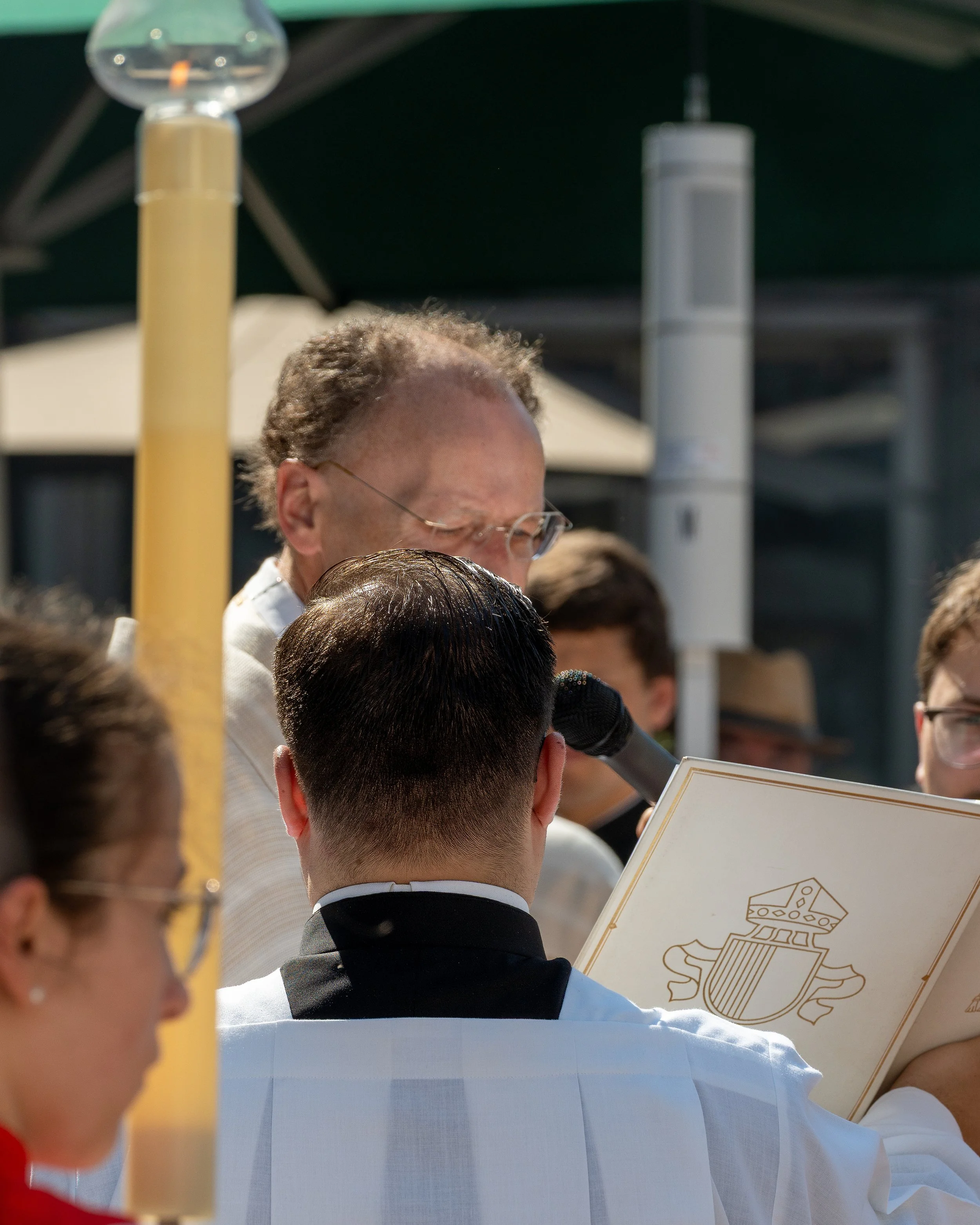 A group of people, including a man with glasses and curly hair, gathered outdoors around a person holding a booklet and speaking into a microphone, with sunny weather and umbrellas in the background.