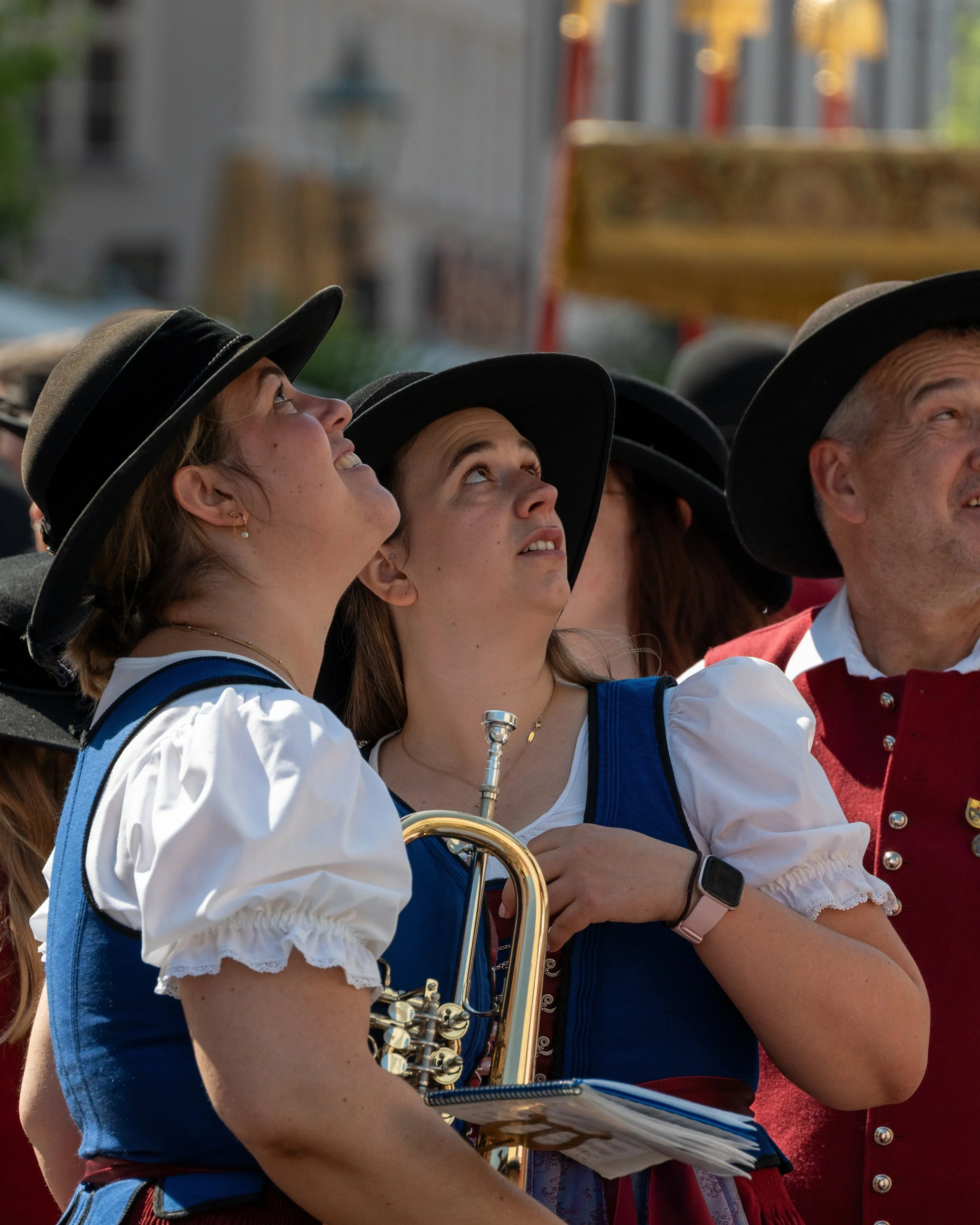 Three women and one man dressed in traditional German attire, looking up and smiling during an outdoor event, with a woman holding a trumpet and a music folder.