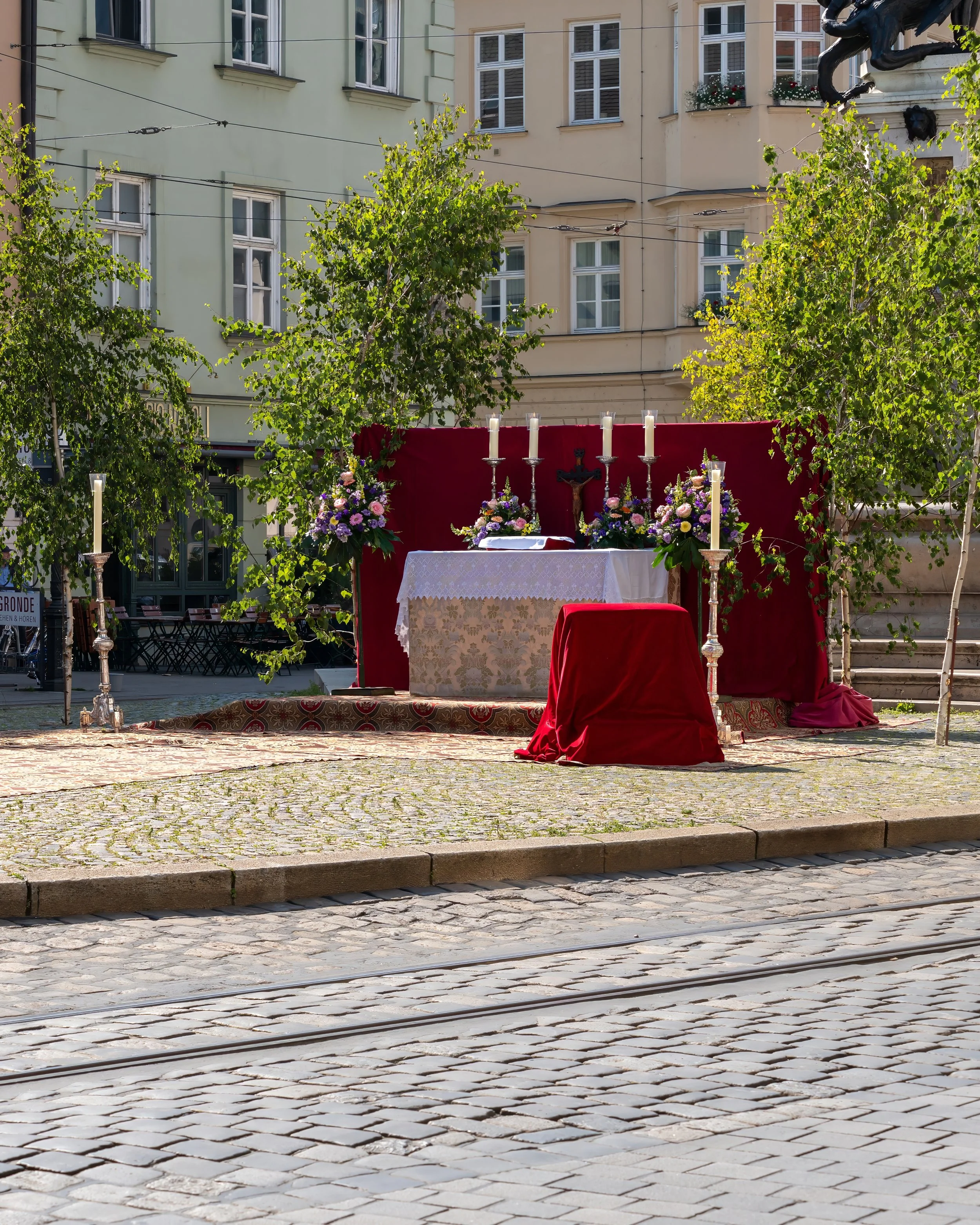 Outdoor altar setup with a priest's chair, candles, flowers, and a crucifix on urban streetscape with trees and buildings in the background.