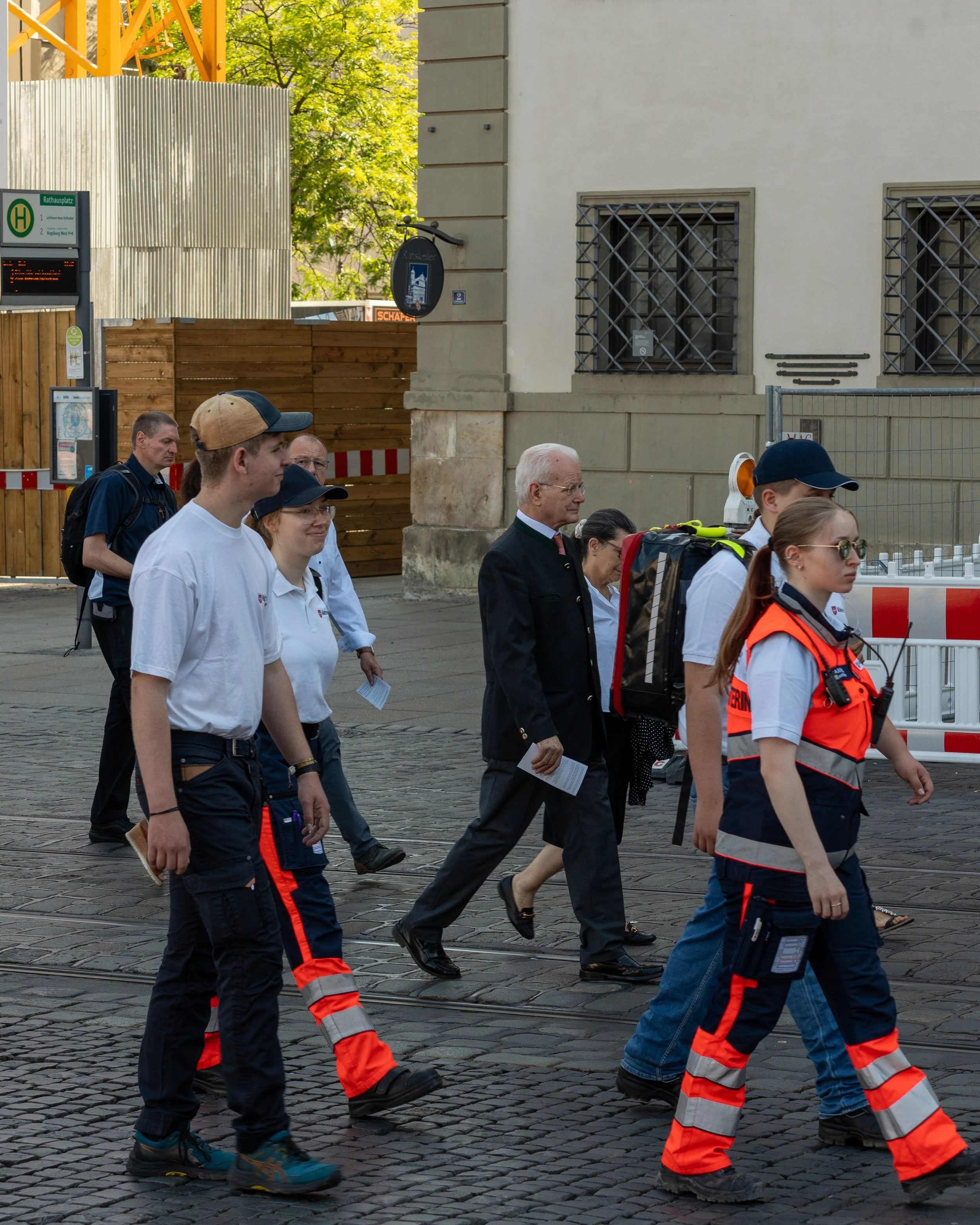Group of people walking on city street, including emergency workers and civilians, with a construction site and building in the background.