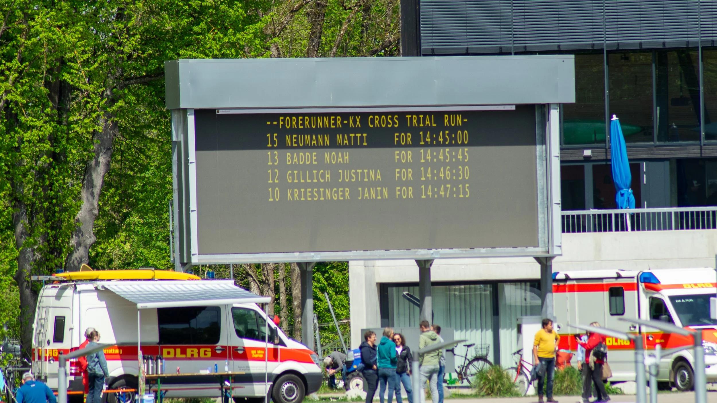 Digital scoreboard at an outdoor sporting event displaying the results of a cross trial run, with participants' names and finish times. Emergency vehicles and people are gathered nearby, with trees and a modern building in the background.