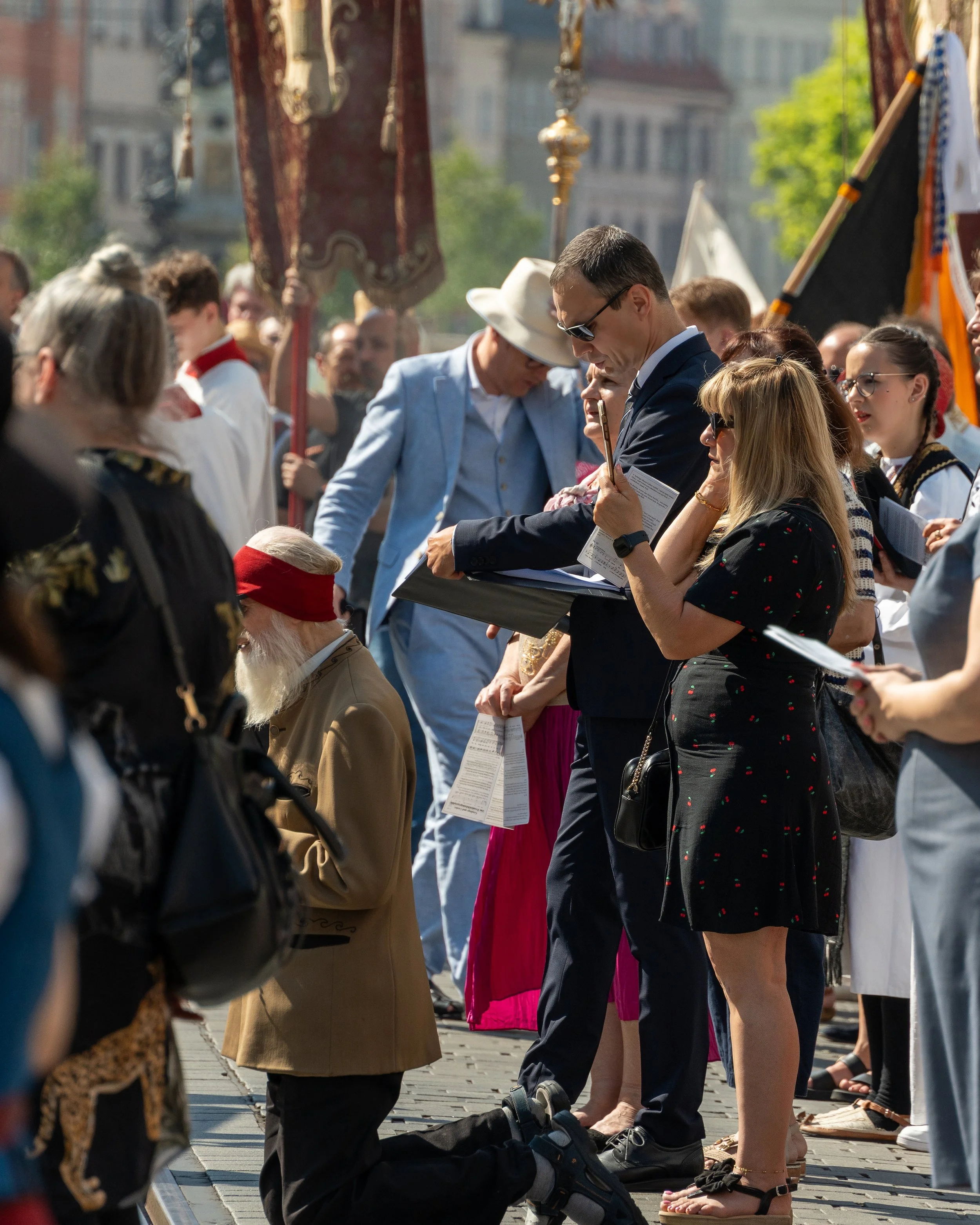 People gathered in a public square, participating in a prayer or moment of silence, with some kneeling and others standing, holding papers, in a sunny outdoor setting with flags and historic buildings in the background.