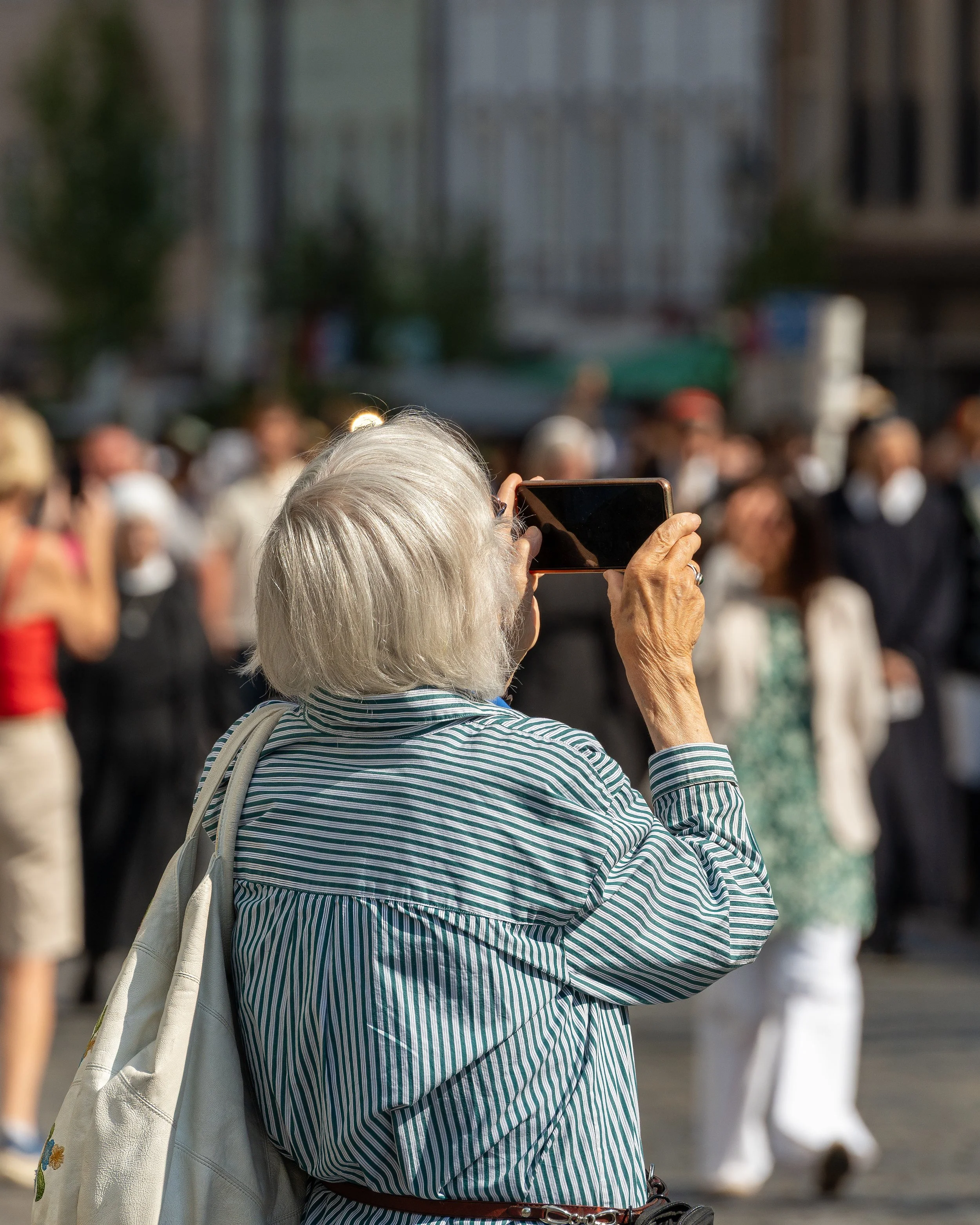 An elderly woman with gray hair taking a photo with her smartphone at a crowd of people in an outdoor urban area.