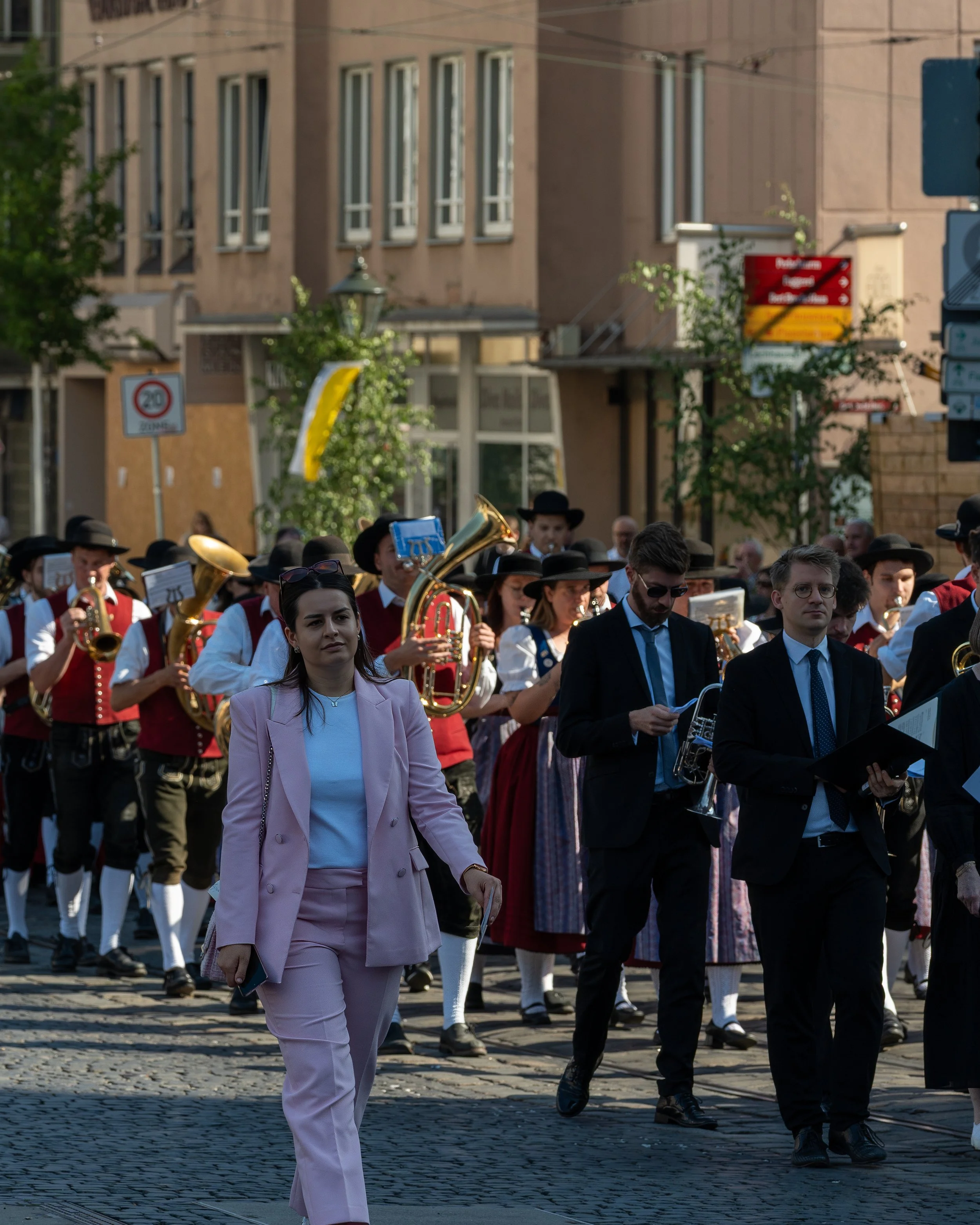 People participating in a street parade, some dressed in traditional clothing and others in formal attire, with a brass band playing music in the background.
