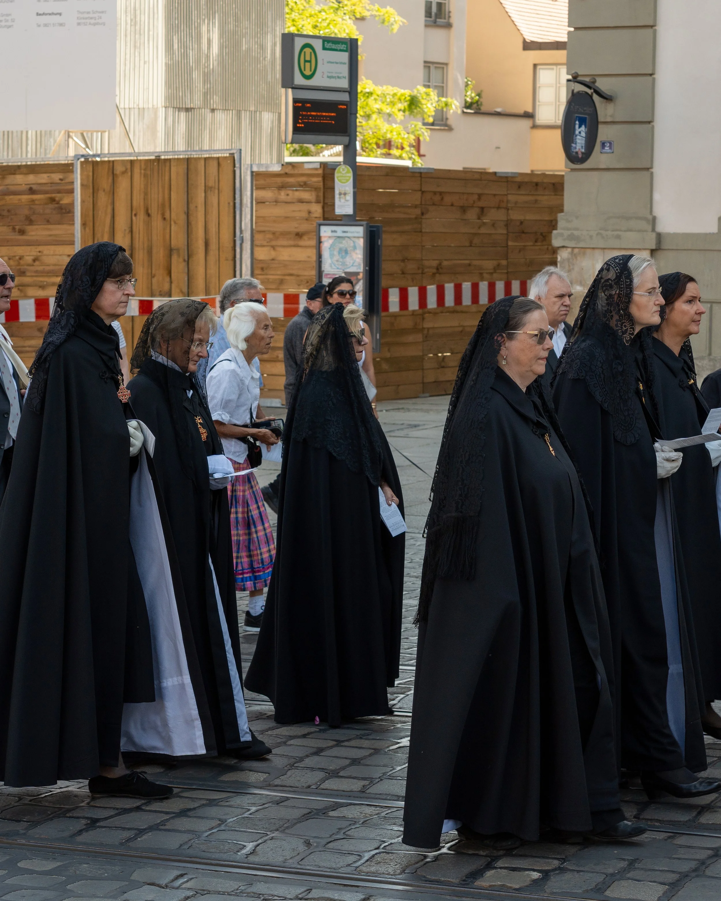 A group of people dressed in traditional black costumes with lace veils participate in a procession on a cobblestone street in an urban area, with a wooden fence, buildings, and a bus stop sign in the background.