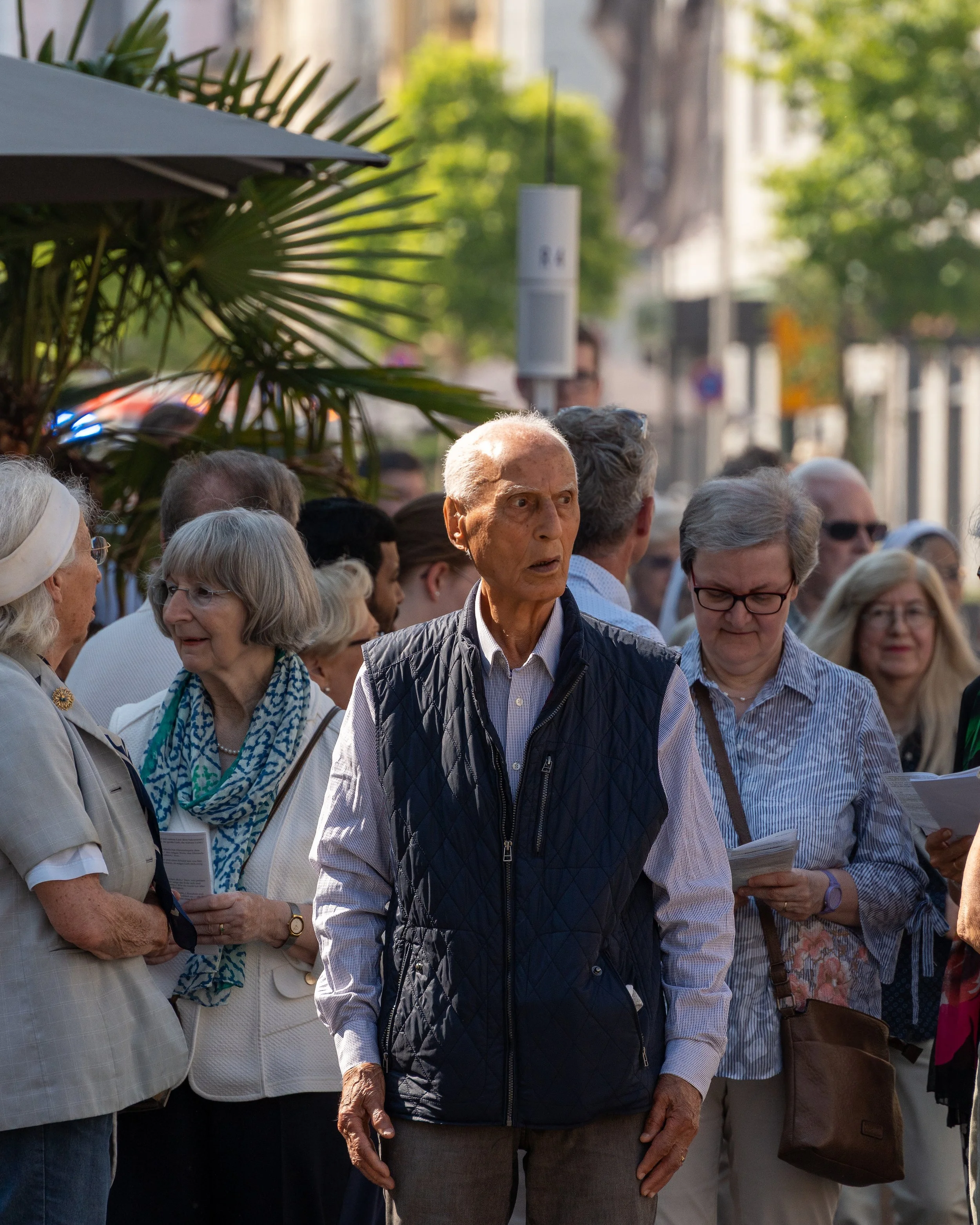 A group of elderly people gathered outdoors in a city street, some holding papers, engaging in a conversation.