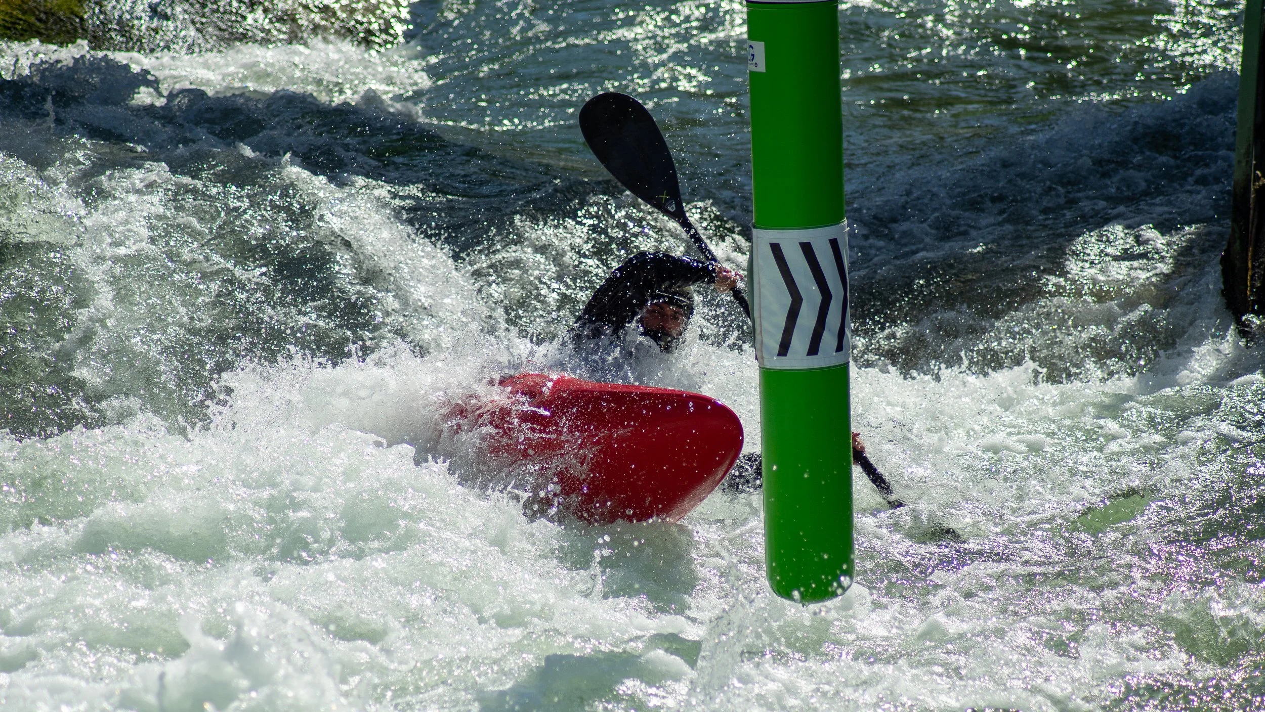 Kayaker in a red kayak navigating through white water rapids, partially obscured by splashing water, with a green and white buoy in the foreground.