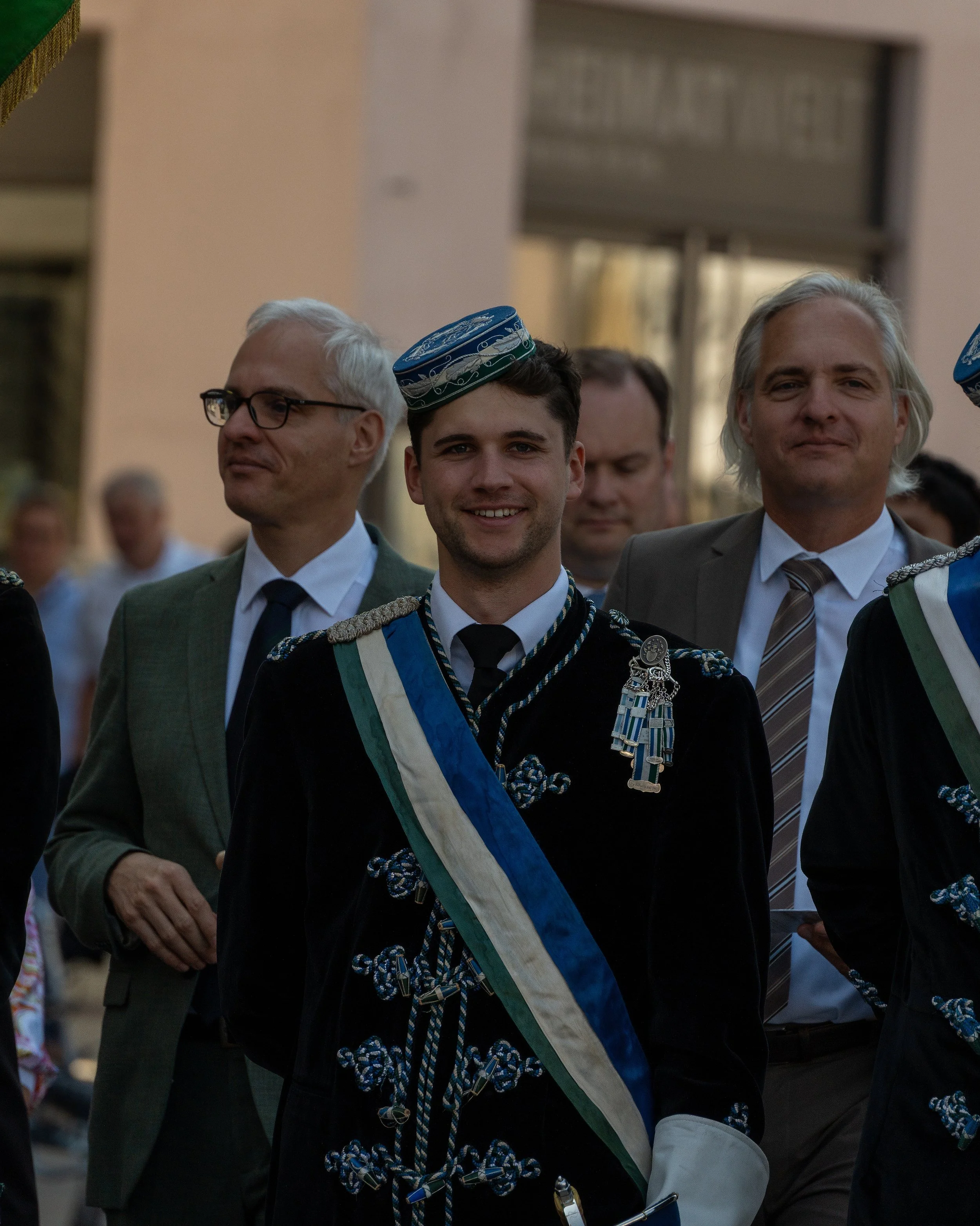 Young man in a ceremonial uniform with a sash and medals, smiling, marching in a parade with other men dressed in suits and traditional attire.