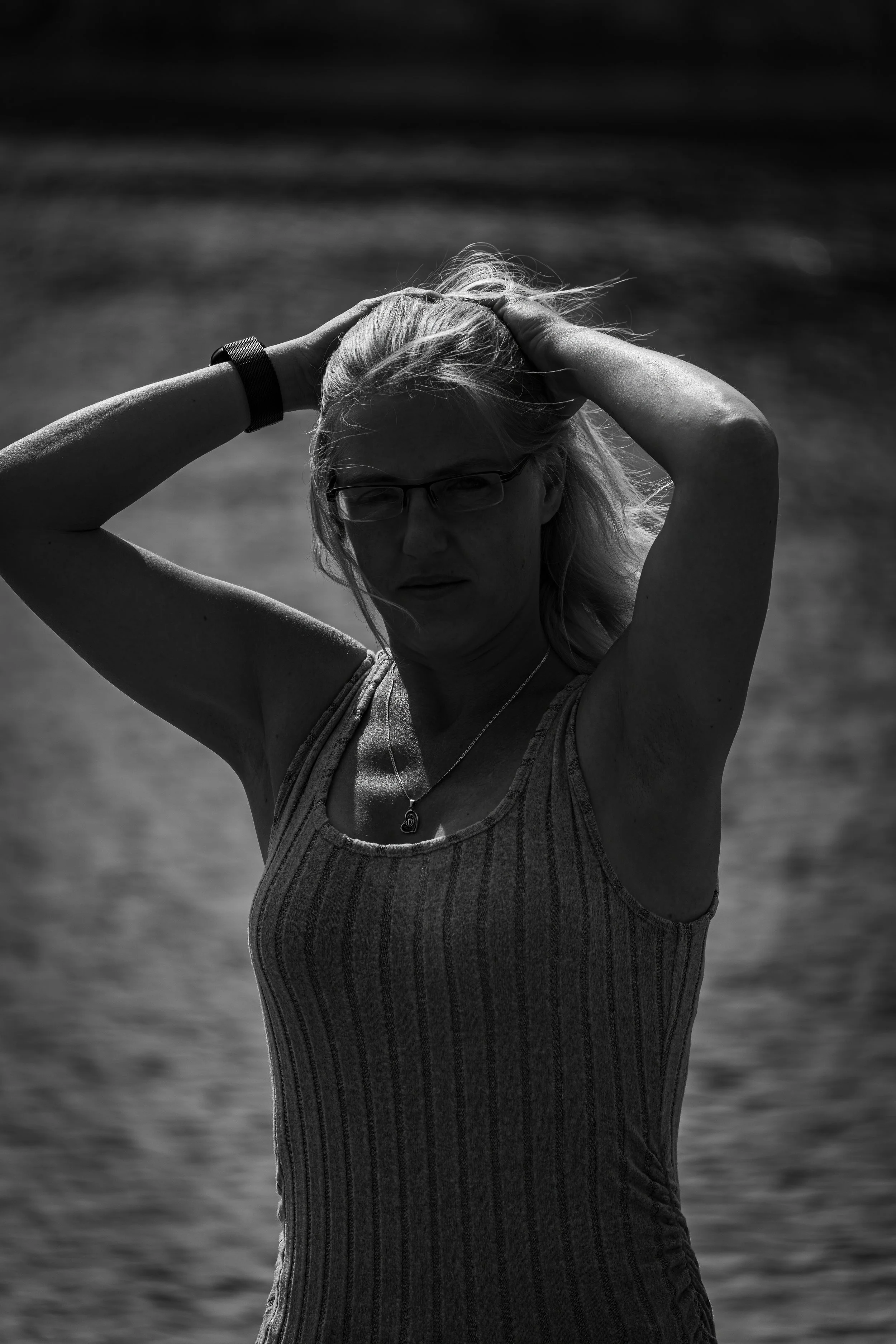 A woman with glasses and a striped tank top standing outdoors near a body of water, with her hands on her head, backlit in black and white