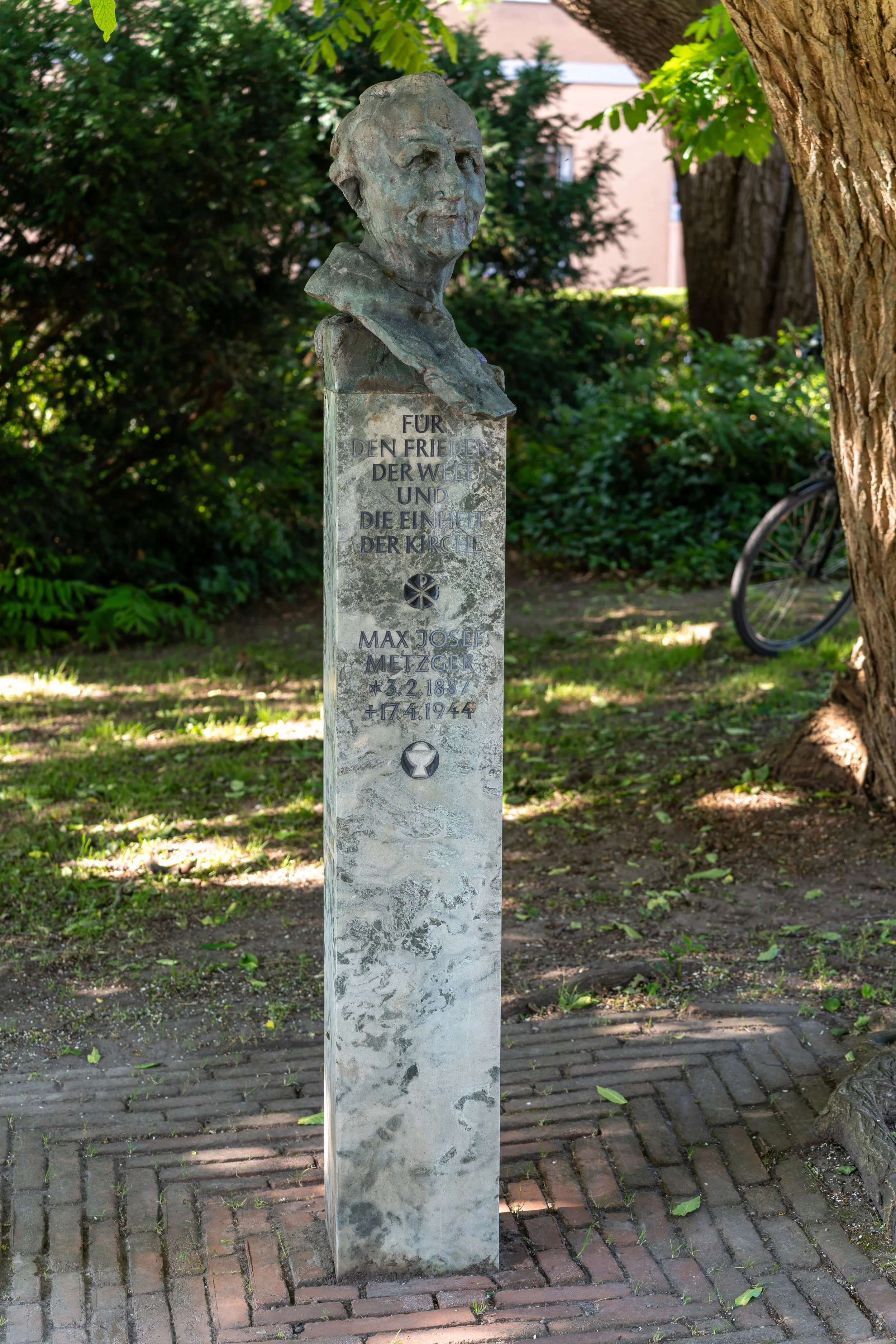 A bust of Max Joseph Metzger on a stone pedestal in a park with green trees and a bicycle in the background.