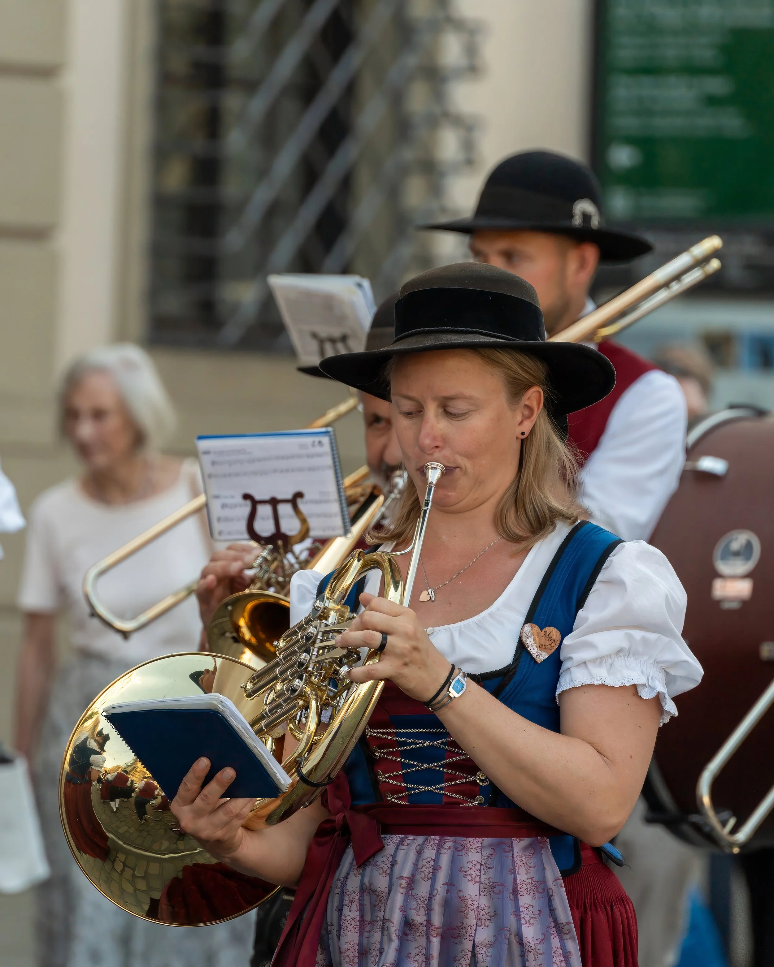 A woman in traditional Bavarian attire playing the saxophone during an outdoor musical performance with other musicians and onlookers in the background.