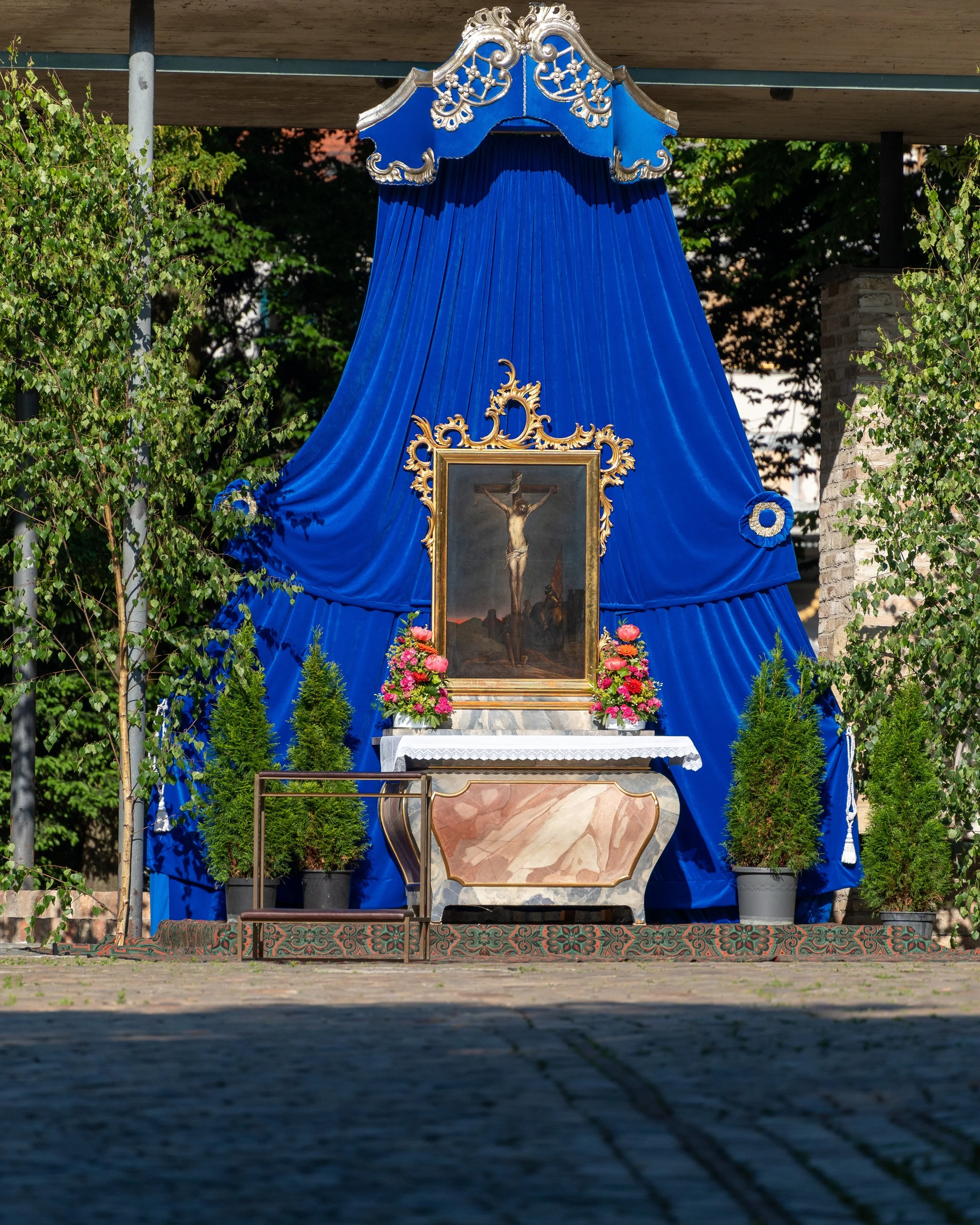An outdoor religious shrine features a framed painting of Jesus on the cross, adorned with pink and white flowers, set on a marble altar with a blue draped fabric backdrop and greenery on the sides.