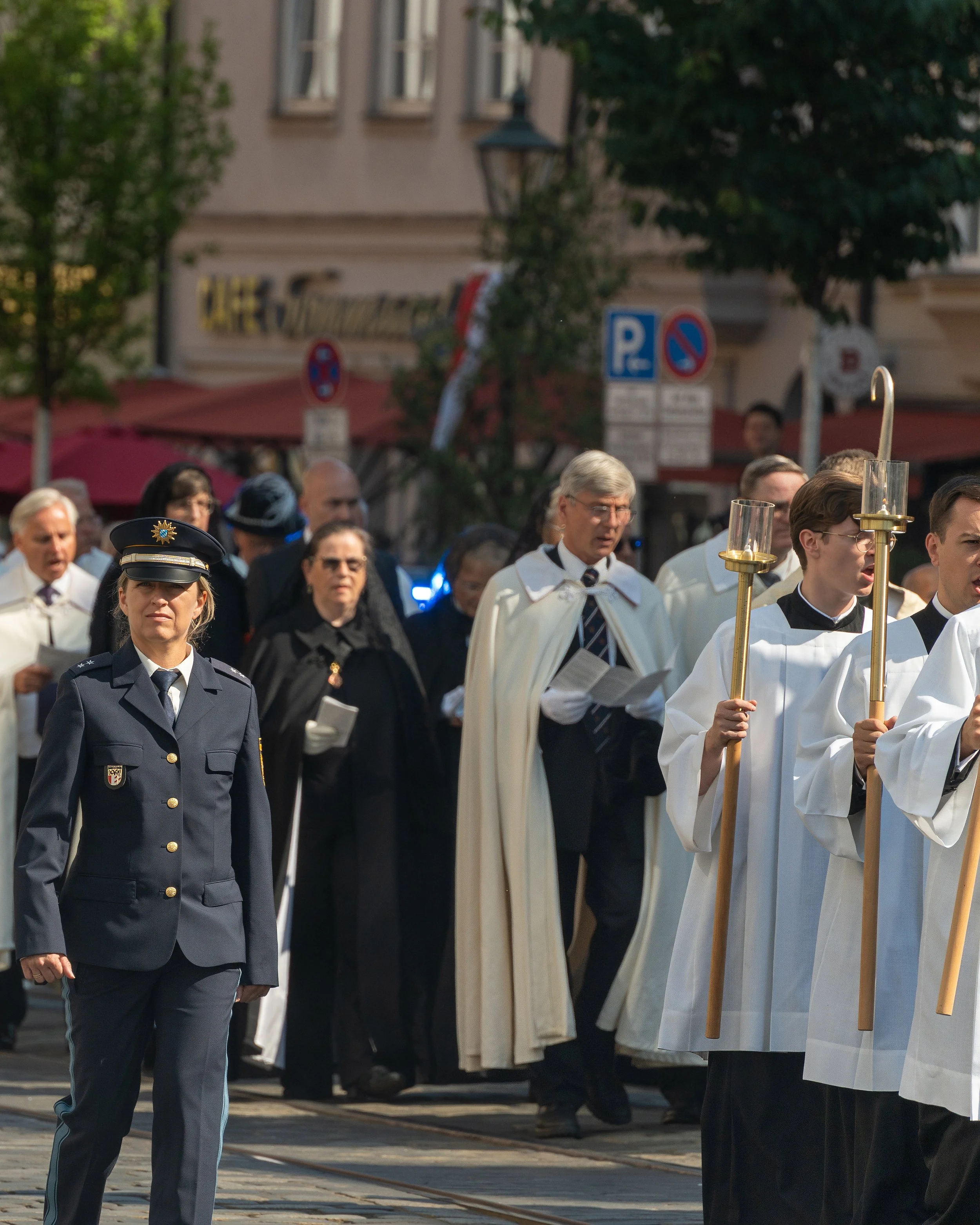 A religious procession with clergy and a police officer walking on a city street, with people in the background, street signs, and storefronts.