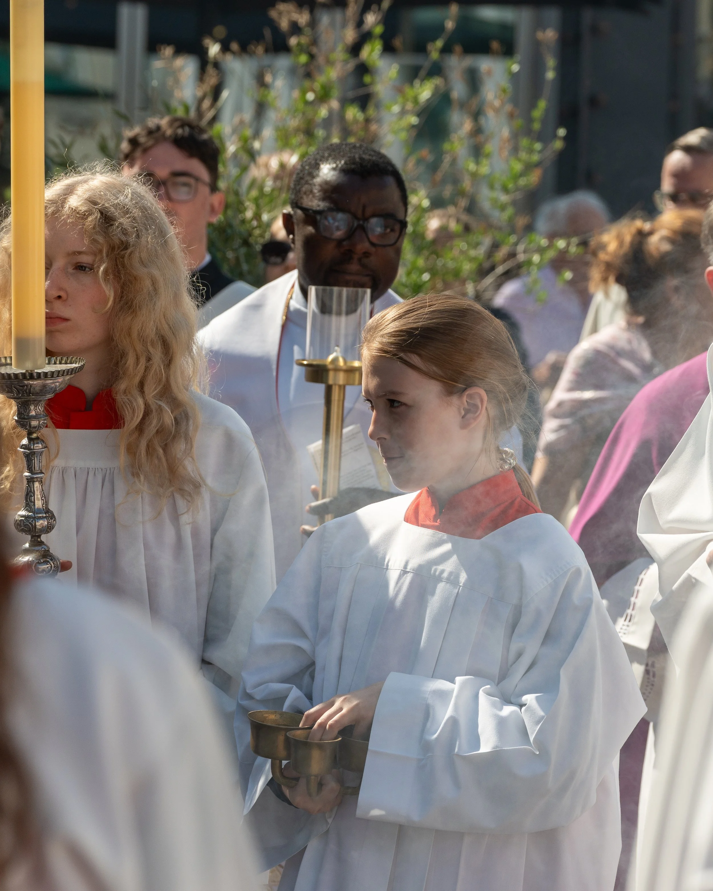 Children in white robes participating in a religious ceremony outdoors, holding candles and trays, with a crowd in the background.