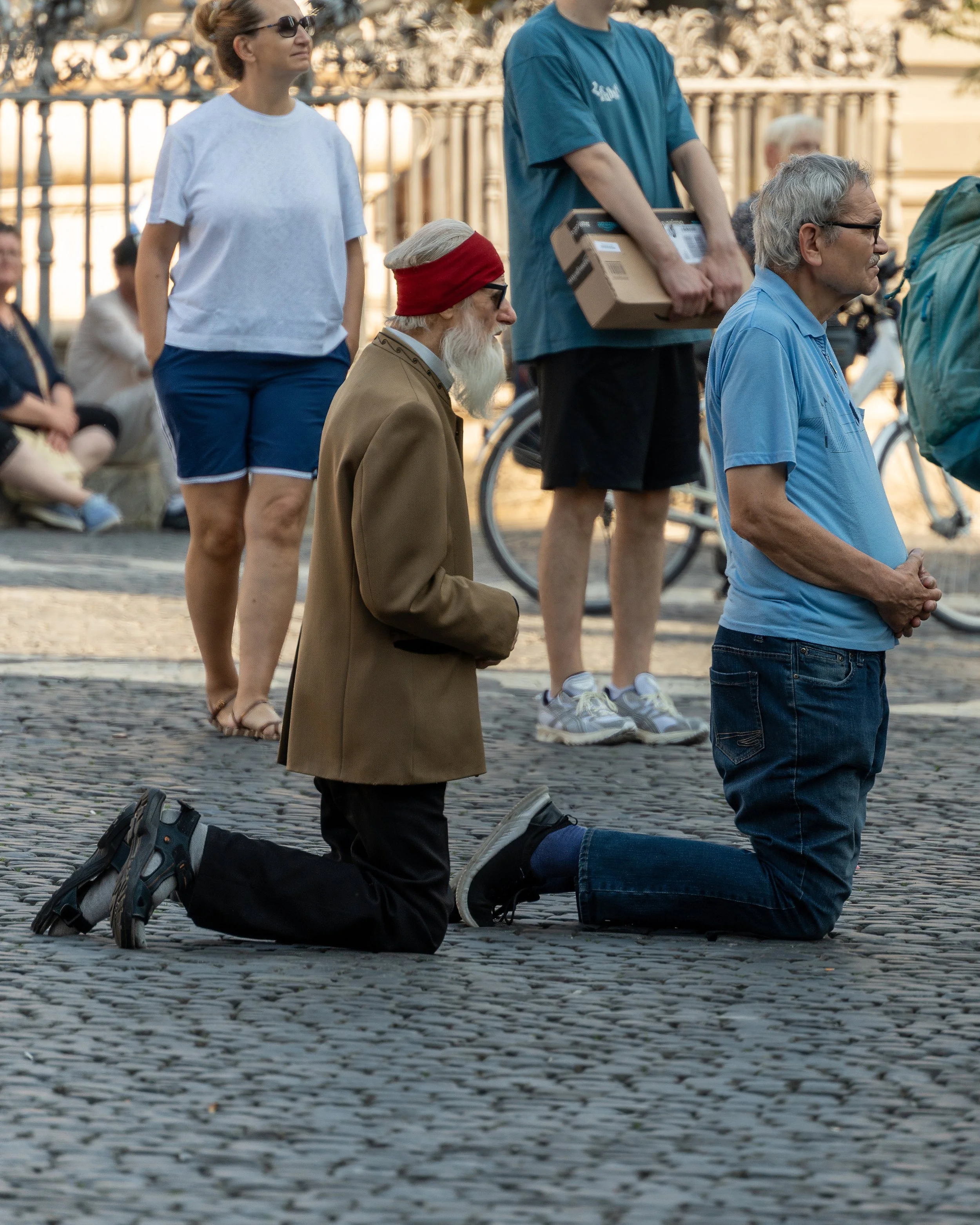 Man in kneeling position with hands clasped, wearing casual clothing, surrounded by protesters, on a cobblestone street.