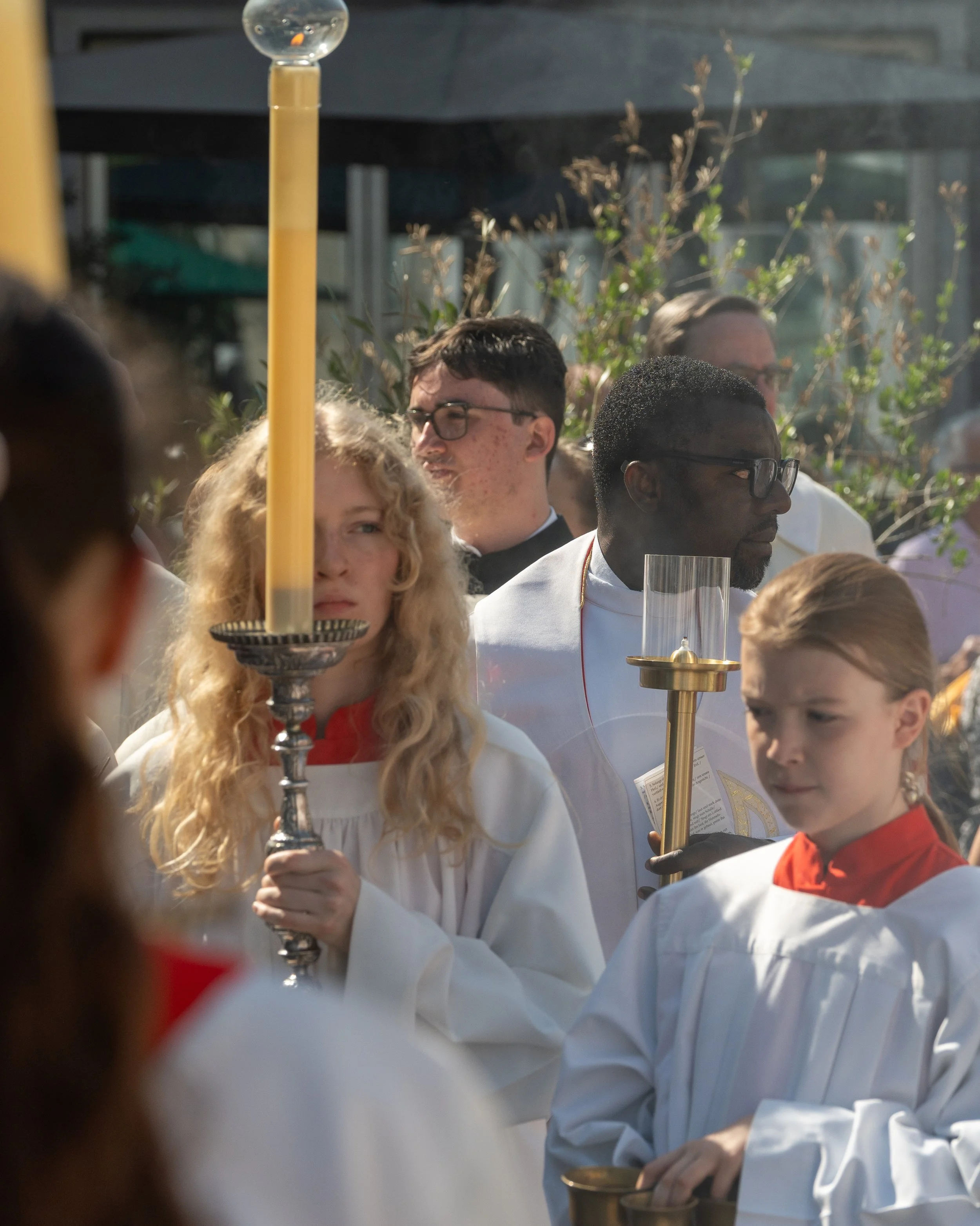 People participating in a religious procession outdoors holding candles and candles in holders, wearing white robes with red collars.