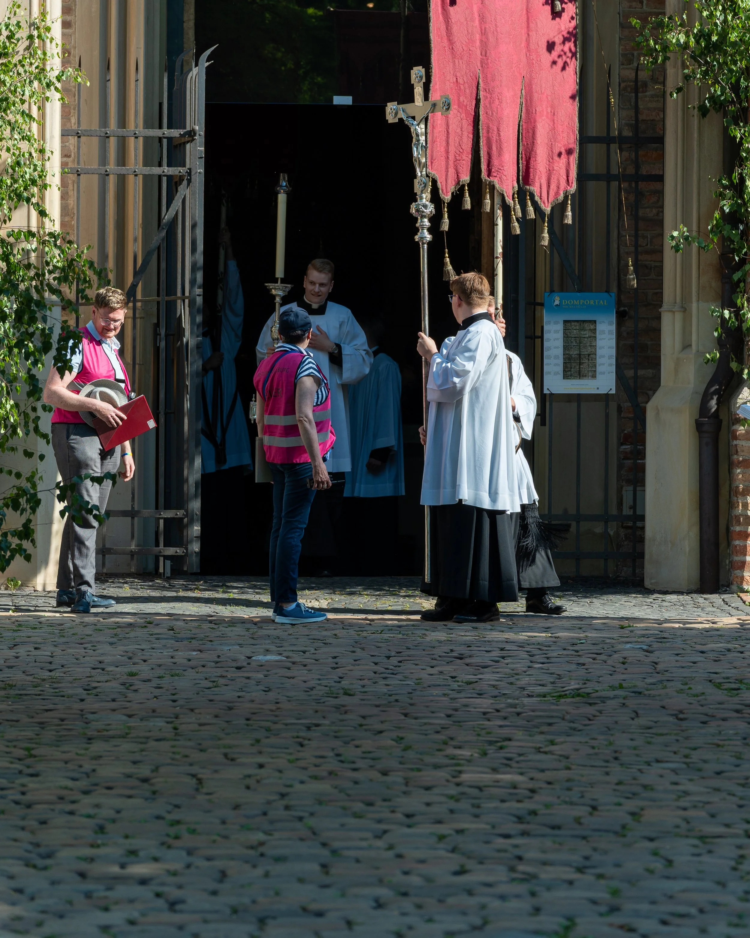 People dressed in clerical robes and laypeople gathered outside a church entrance, participating in a religious procession. Some hold candles, a cross, and a canopy, as part of a religious ceremony.