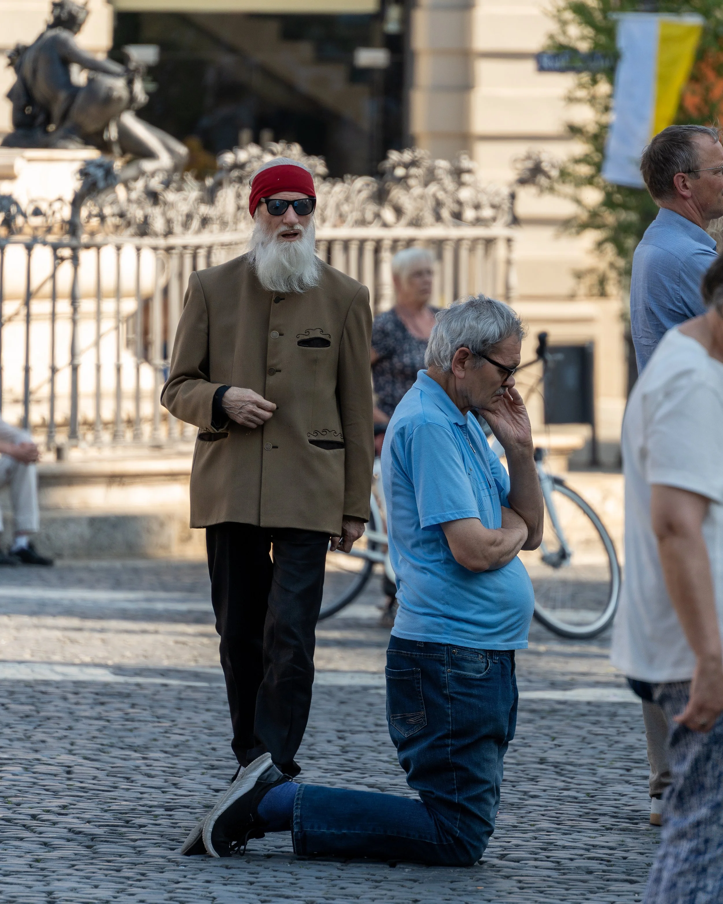 A man kneeling on the cobblestone street with his hand on his face, and an older man standing nearby with a surprised expression wearing a red hat and sunglasses. Other individuals are in the background, suggesting a public gathering.