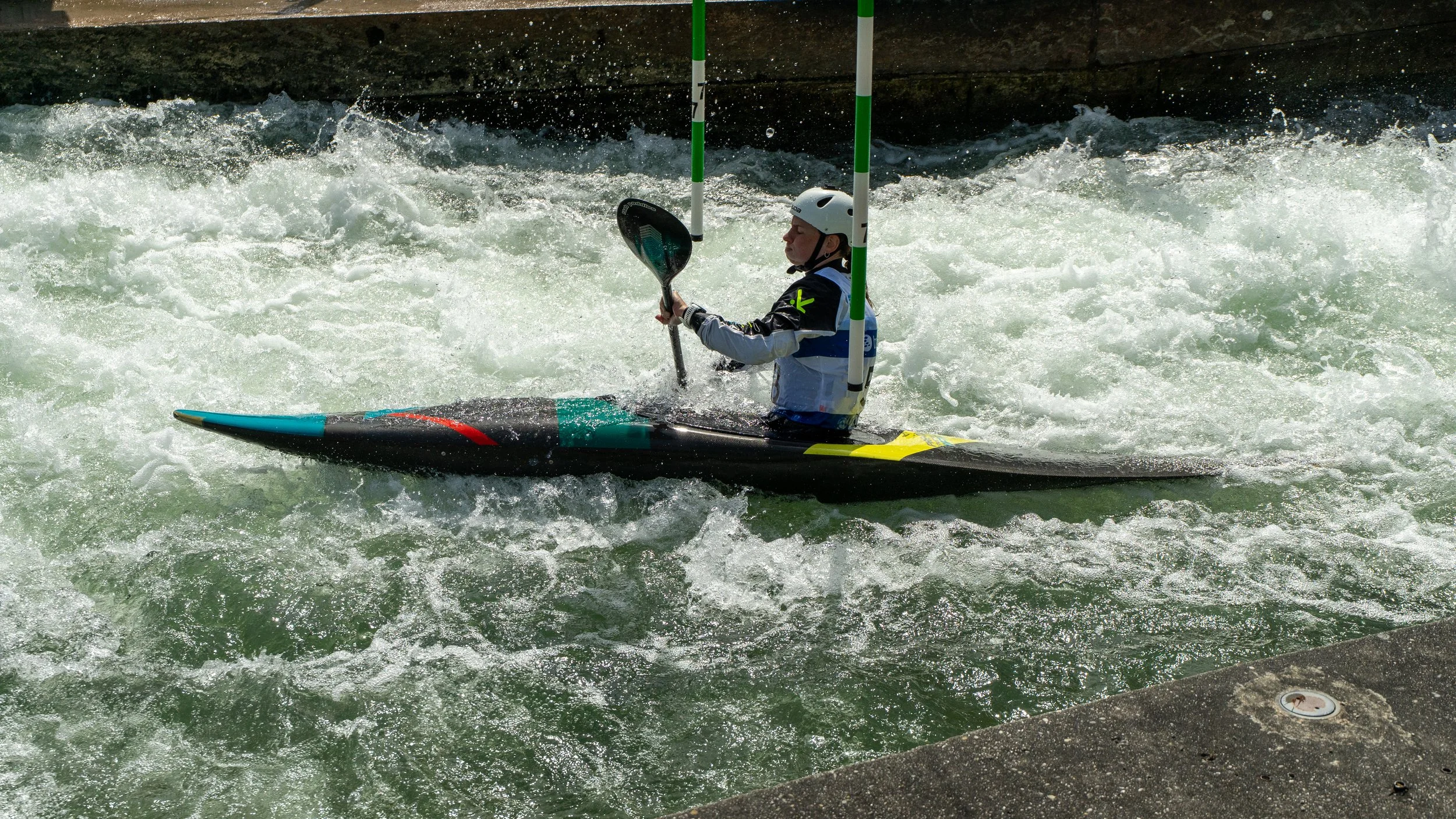 Kayaker navigating a slalom course in rapid white water, holding a paddle under two hanging slalom poles.