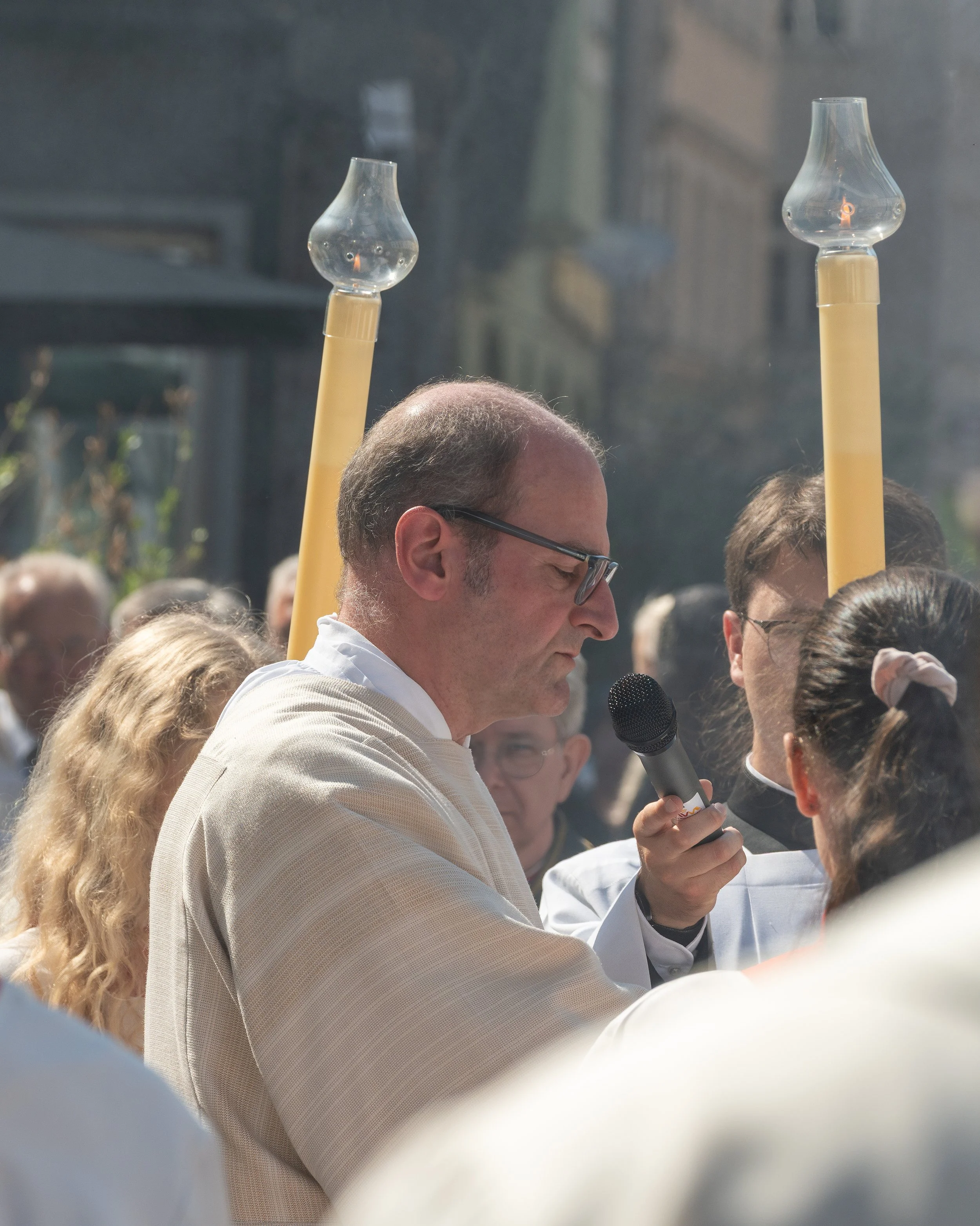 A man in glasses holding a microphone during a large outdoor gathering.