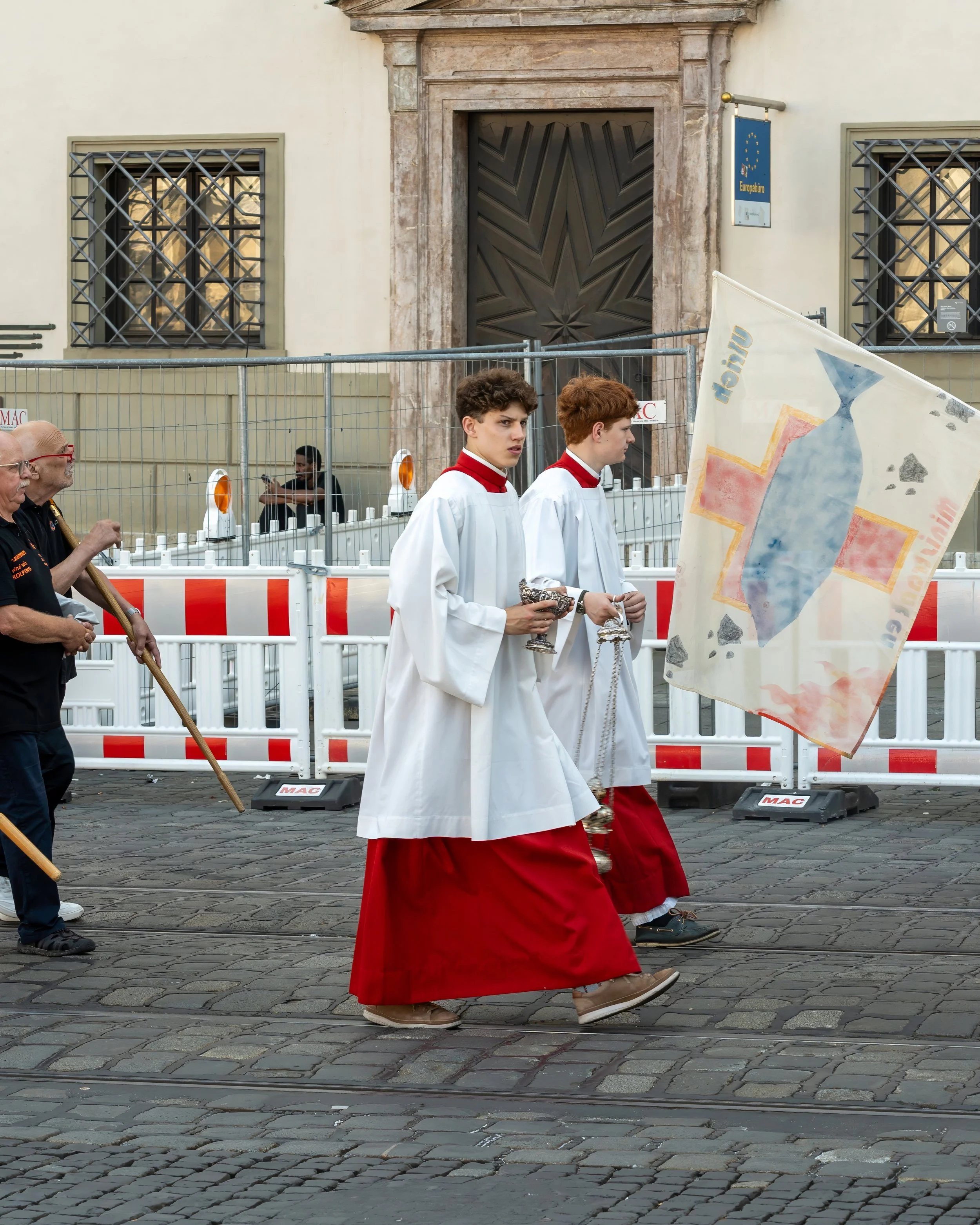 Two young men dressed as altar servers in white surplices with red cassocks walk on a cobblestone street during a parade, carrying a silver chalice and a container, with a banner featuring a stylized bottle and cross in the background. Behind them, a
