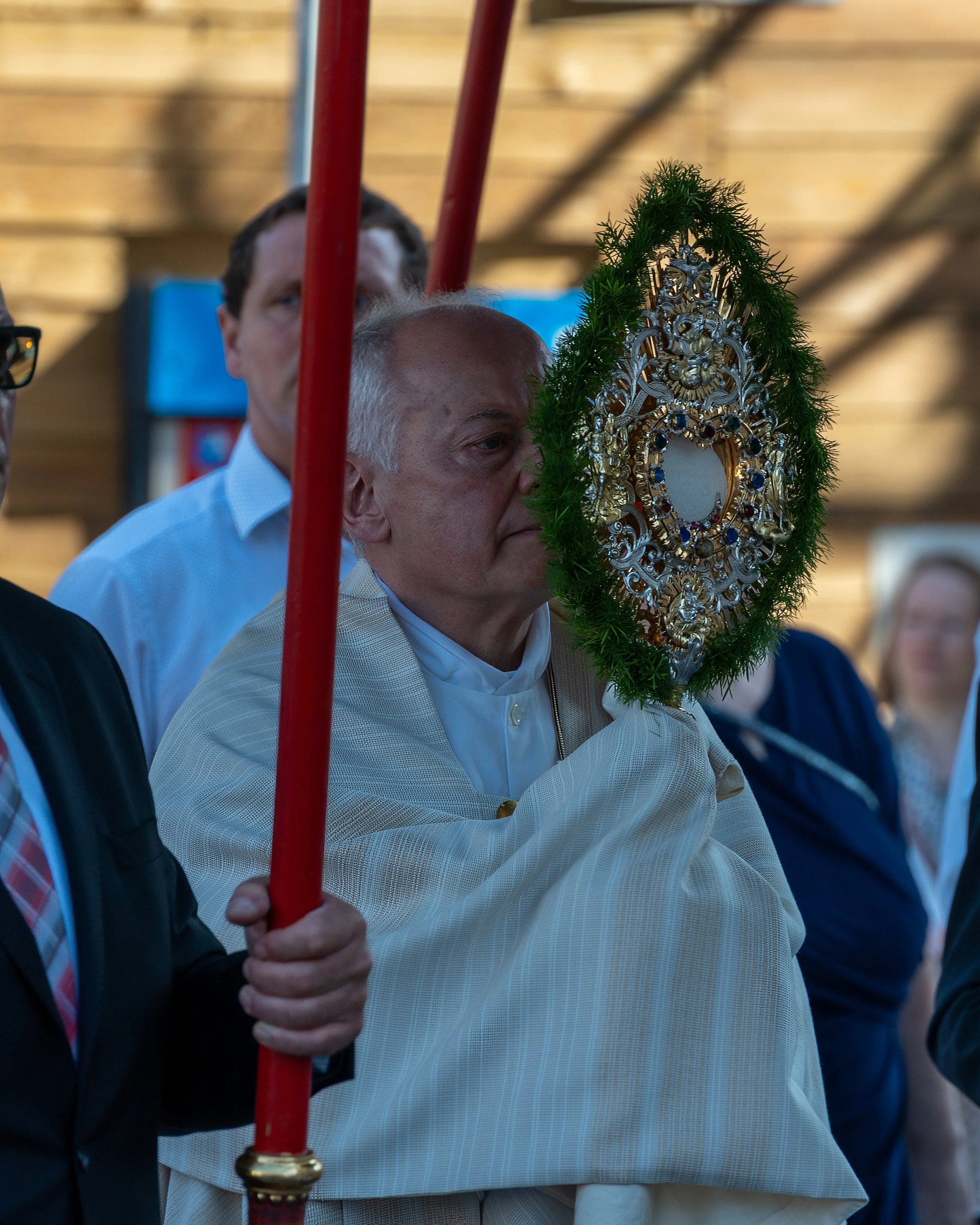 A religious procession with a priest holding an ornate silver and gemstone monstrance, partially covered by a decorative frame, while others stand around him.
